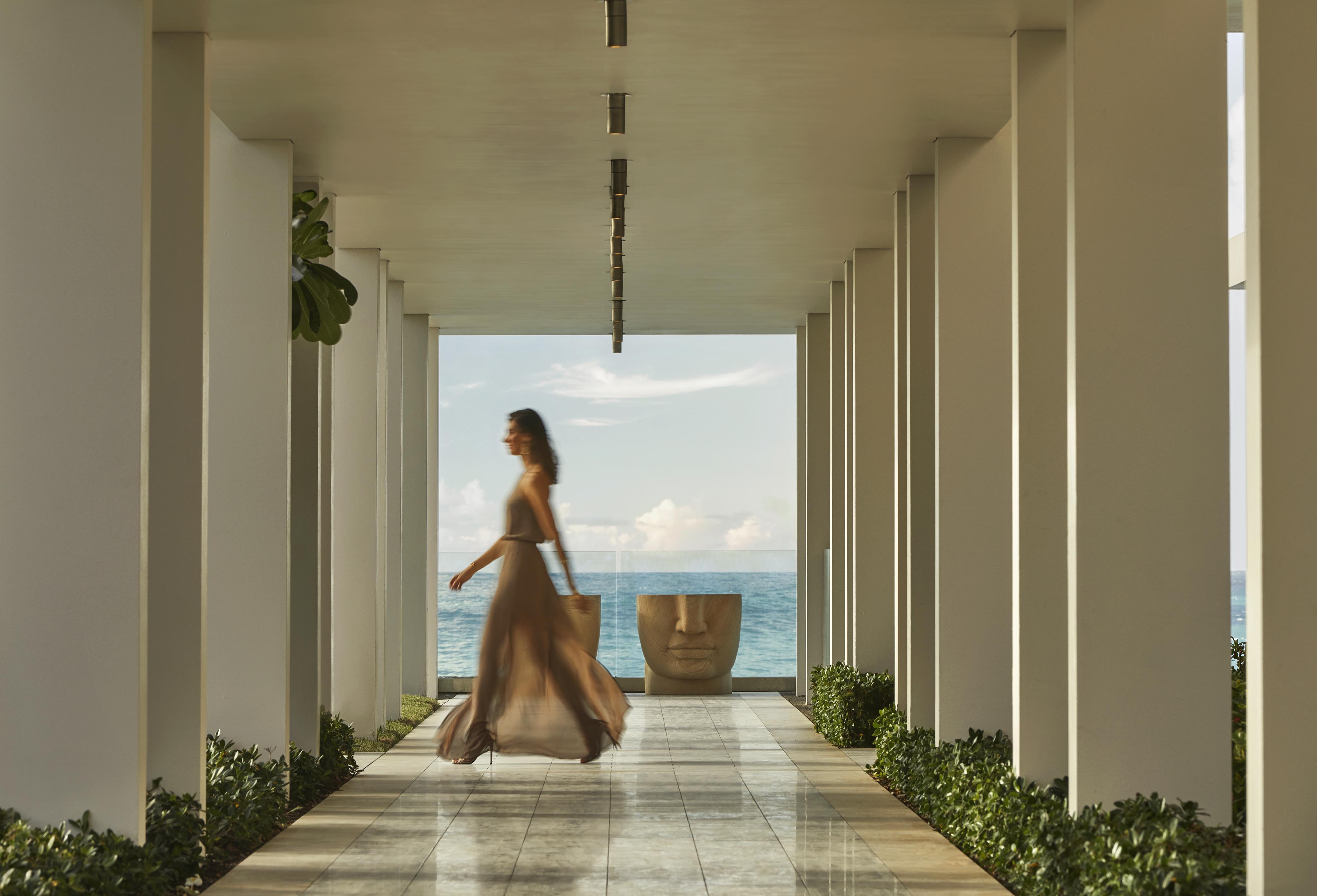A person in a flowing dress walks down a corridor with white pillars, leading to a large, face-like sculpture. Ocean view is visible in the background.
