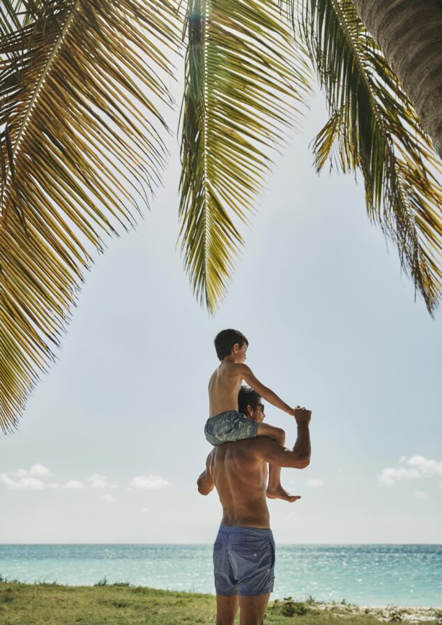 A shirtless man carries a child on his shoulders near the ocean, surrounded by palm trees, with a clear sky above.