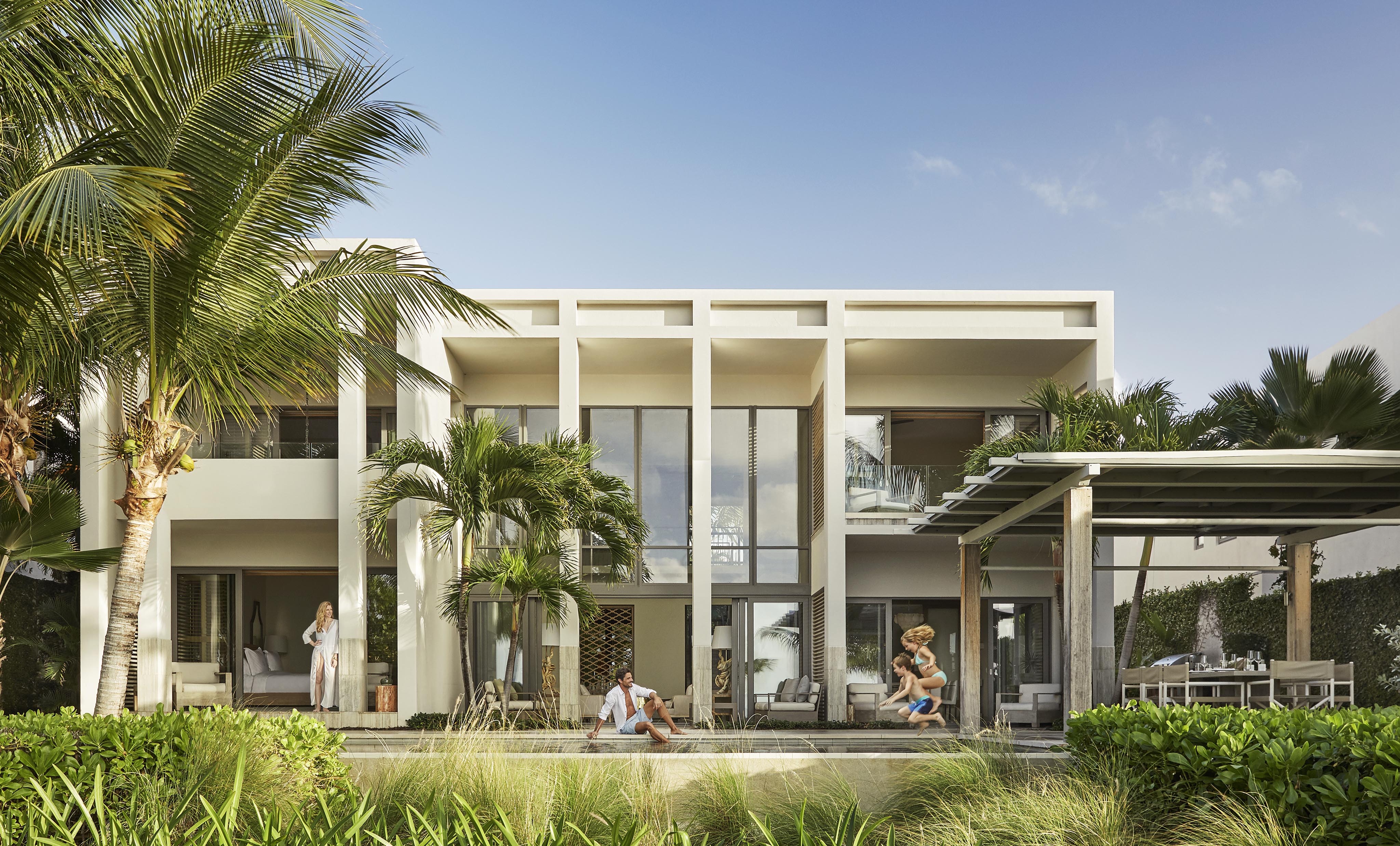 Modern two-story house with large windows, surrounded by palm trees. People are relaxing outside on a sunny day with patio seating and a dining area.