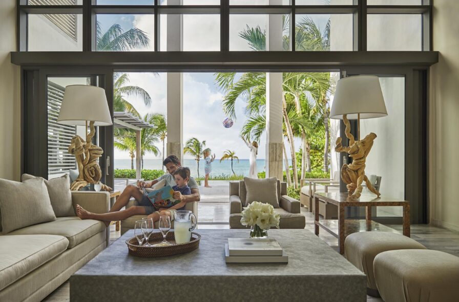 A person reads a book with a child on a sofa in a modern living room. Large windows display a tropical outdoor view with palm trees and a pool. Elegant decor and furnishings complete the scene.