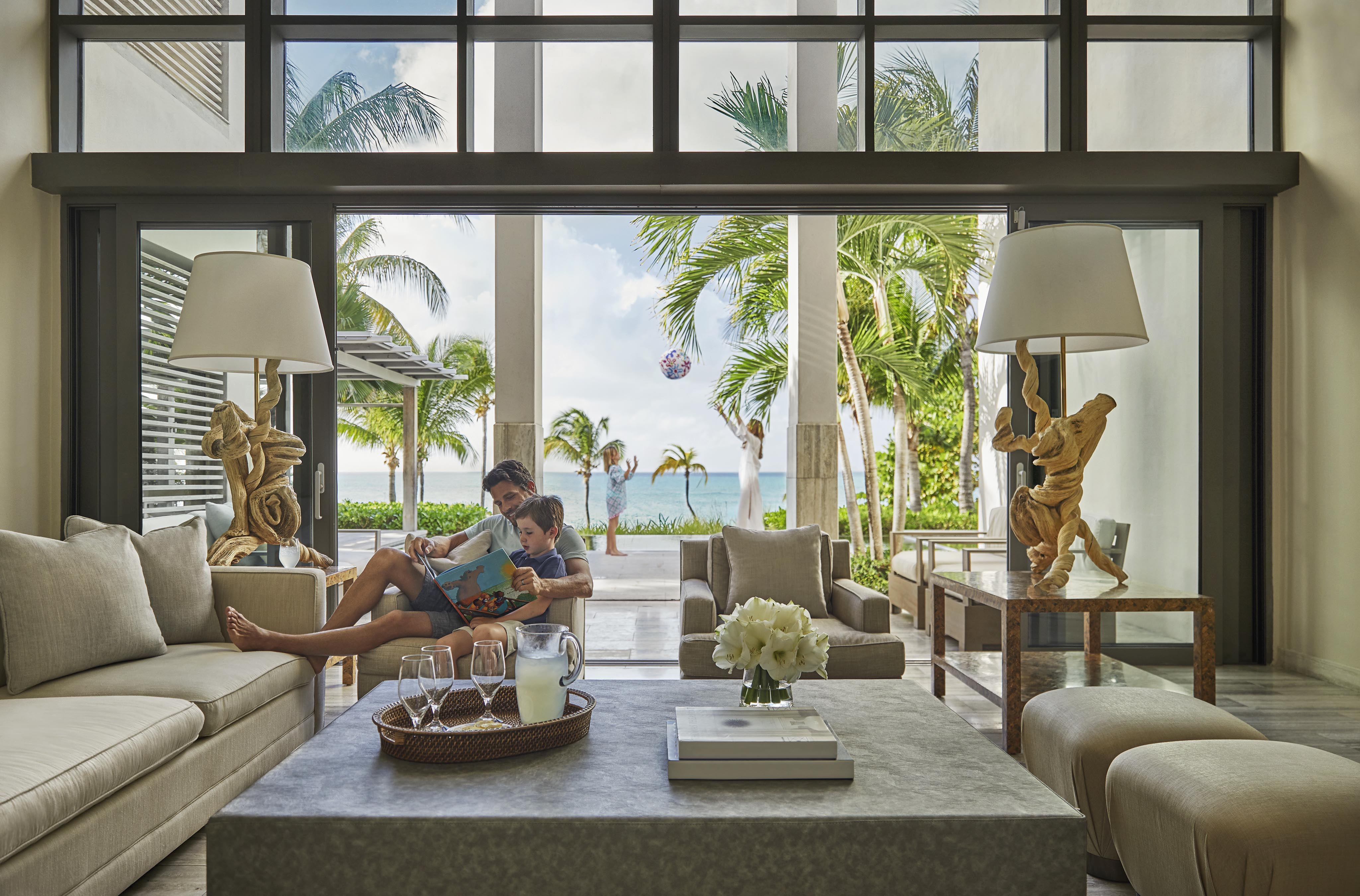 A person reads a book with a child on a sofa in a modern living room. Large windows display a tropical outdoor view with palm trees and a pool. Elegant decor and furnishings complete the scene.