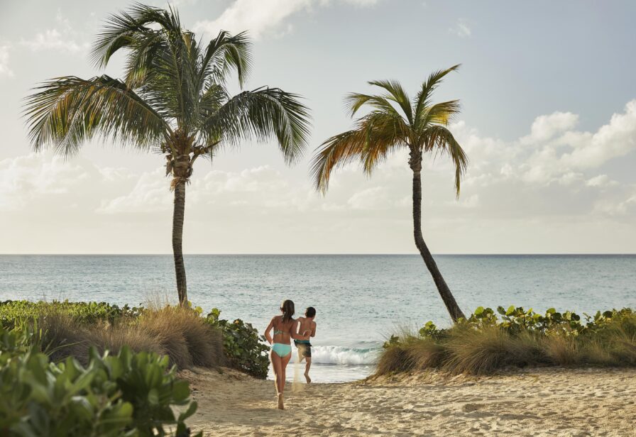 Two people in swimwear walk towards the ocean on a sandy beach, framed by two palm trees, with a partly cloudy sky above.