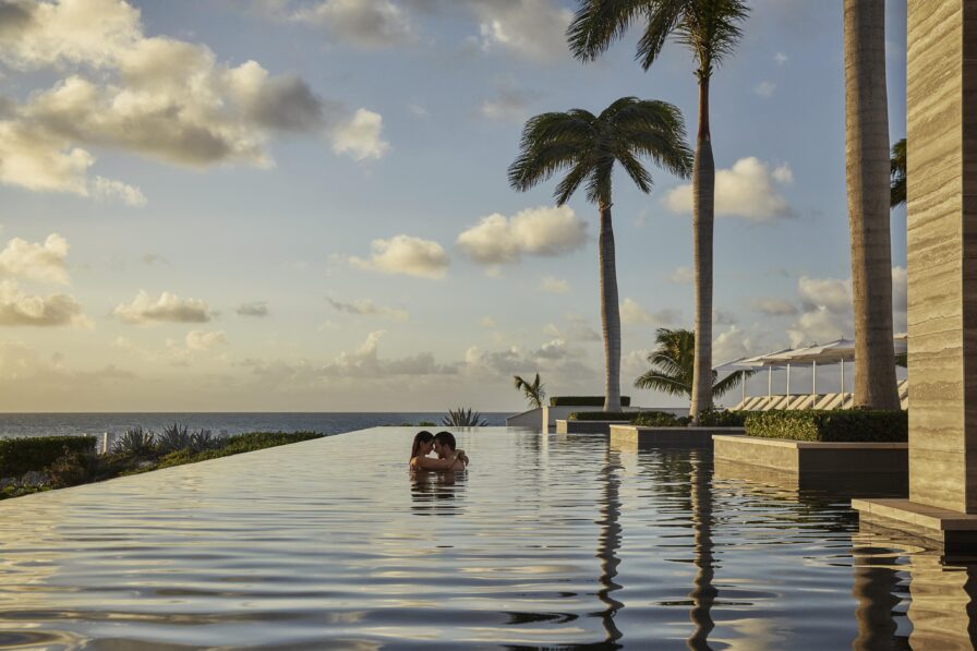 Two people are in an infinity pool overlooking the ocean with scattered clouds in the sky and palm trees nearby.