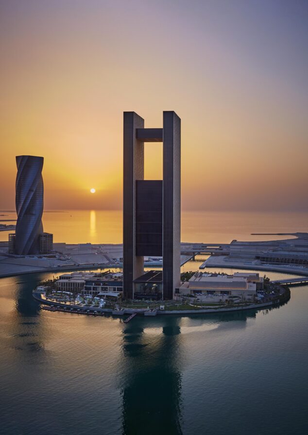 A tall, modern Bahrain skyscraper with a rectangular cutout stands on an island at sunset, surrounded by water and adjacent buildings.