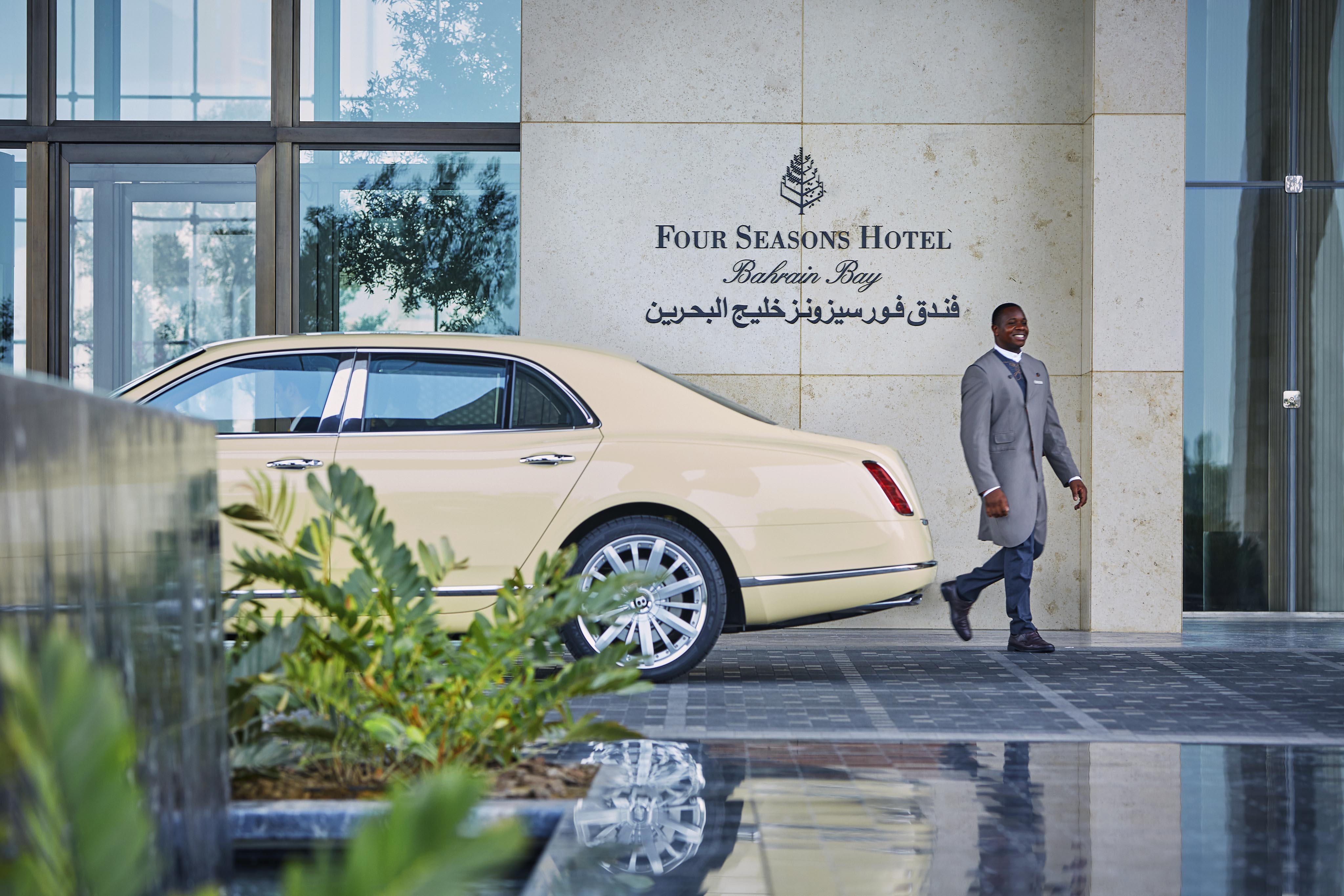 A man in a gray suit walks past a parked beige luxury car outside the entrance of Four Seasons Hotel Bahrain Bay.