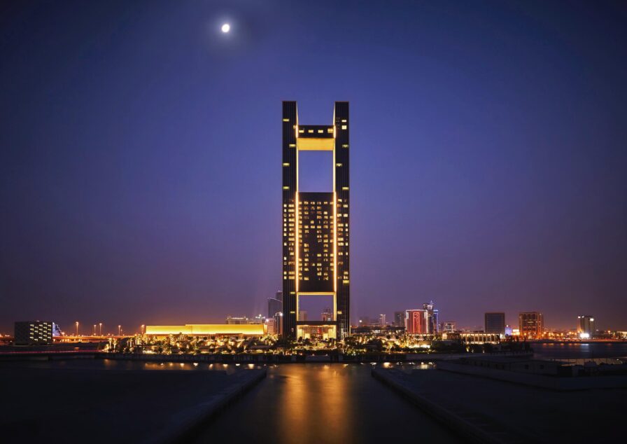 An aerial architectural view of Four Seasons Bahrain exterior ; a towering rectangular skyscraper with illuminated windows standing against a night sky with a visible moon, surrounded by city lights and lower buildings.