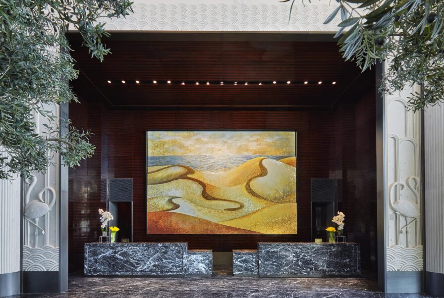 A luxury hotel lobby interior of Four Seasons Bahrain Bay features two black marble reception desks, potted plants, and a large abstract painting of sand dunes on a dark wood wall—perfect for resort photography in the Middle East.