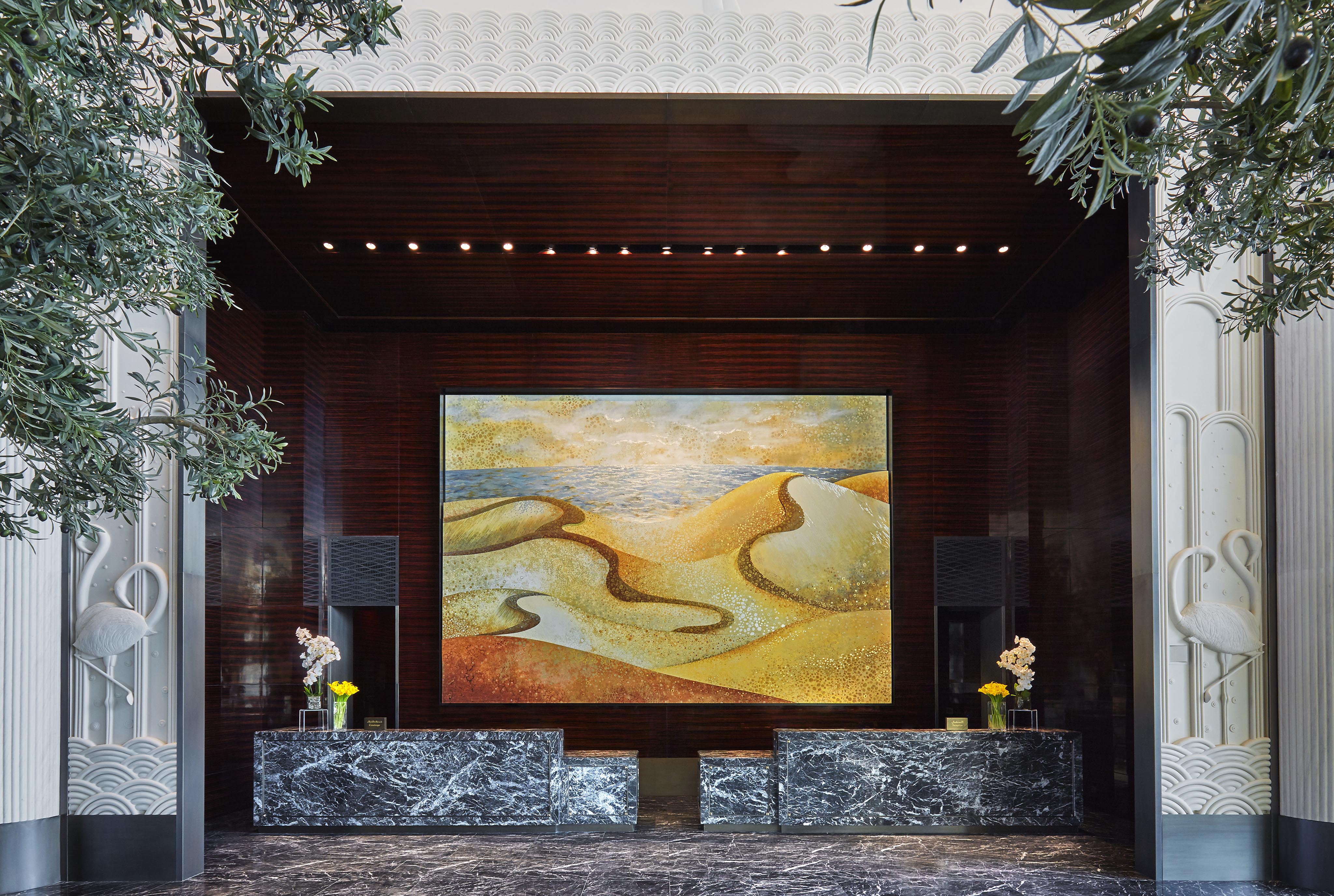 A luxury hotel lobby interior of Four Seasons Bahrain Bay features two black marble reception desks, potted plants, and a large abstract painting of sand dunes on a dark wood wall—perfect for resort photography in the Middle East.
