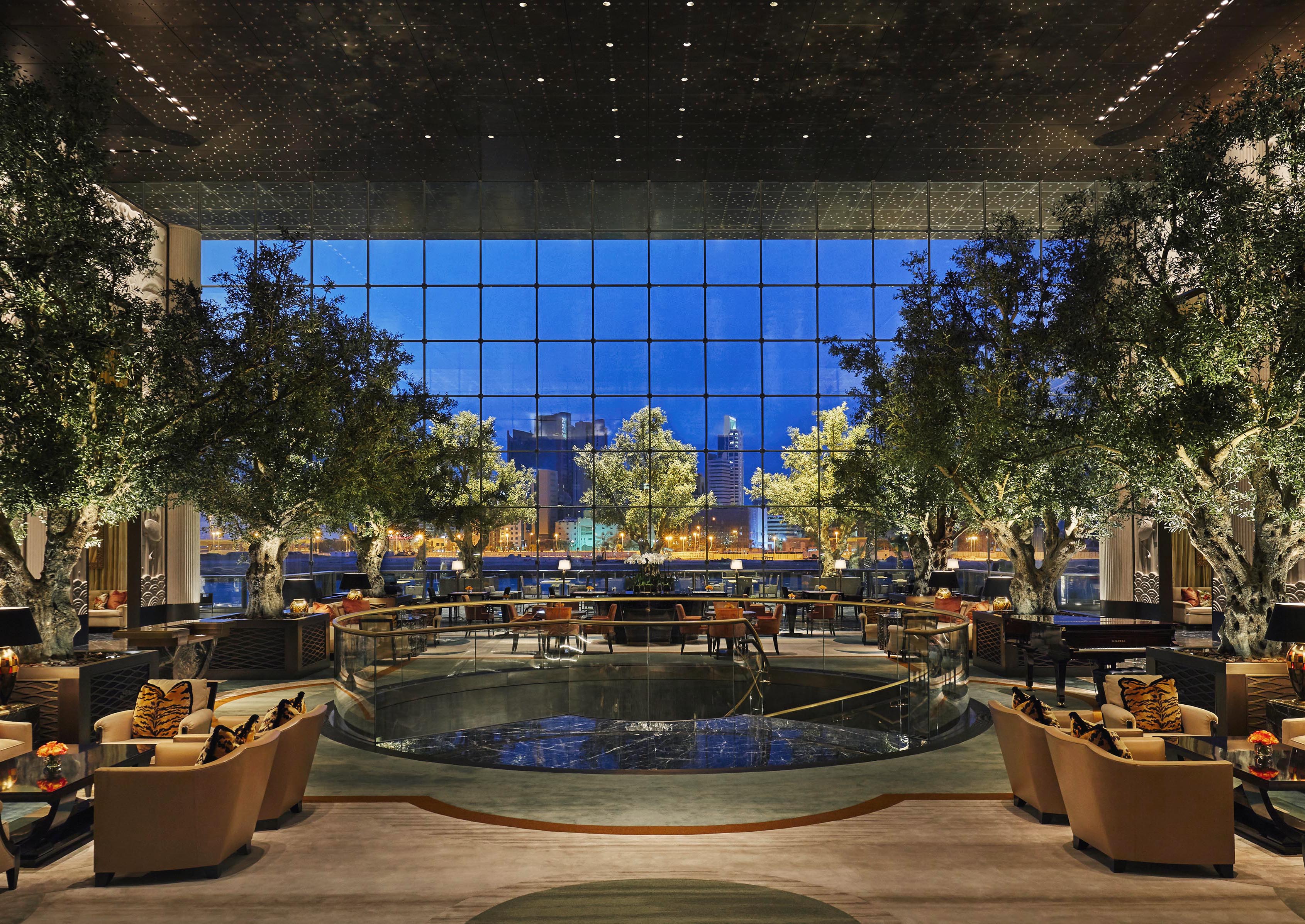 A spacious Four Seasons Bahrain hotel lobby with luxury modern seating, tall indoor trees, and large windows offering a cityscape view at dusk.
