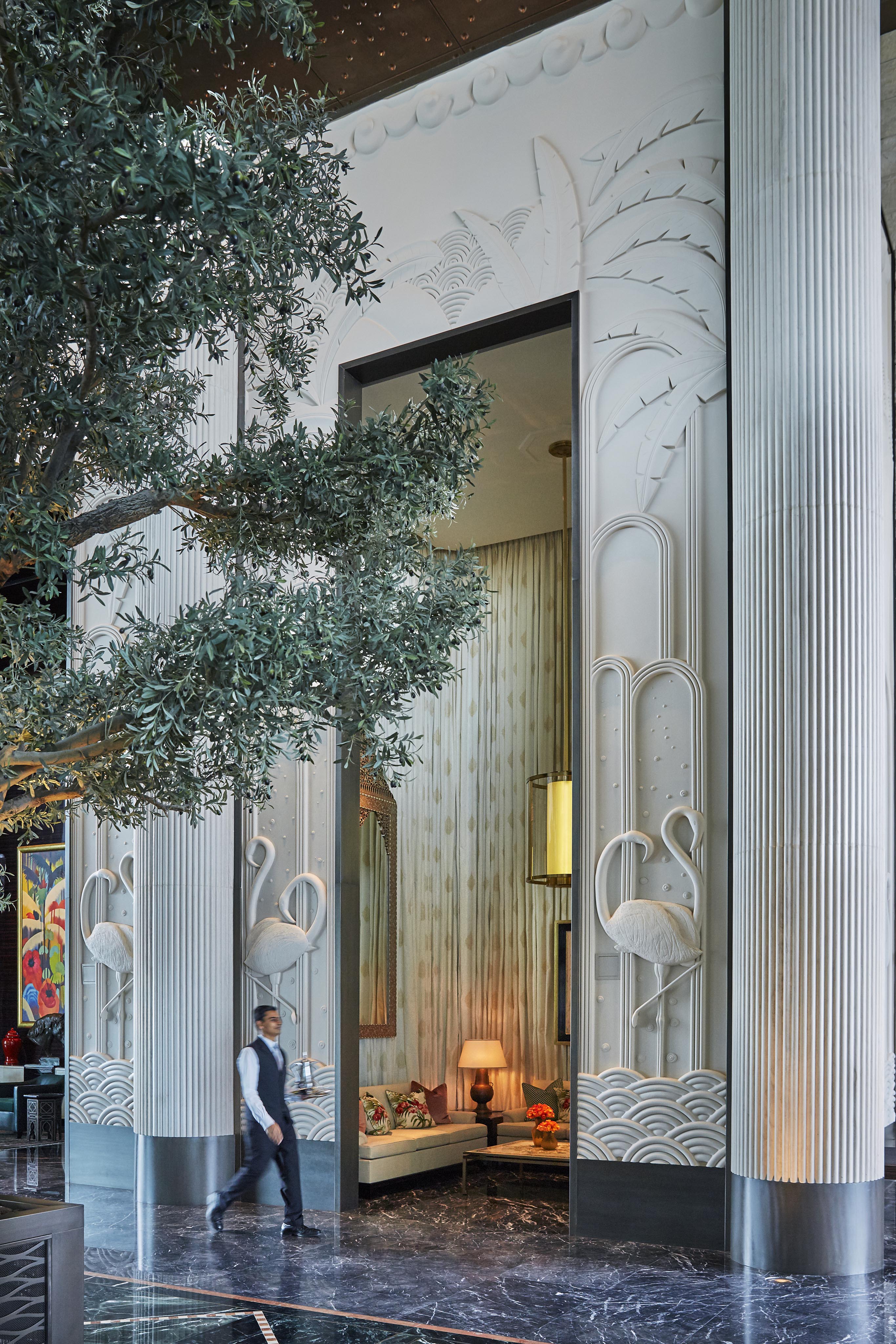 A man in uniform walks through an ornate Four Seasons Bahrain Bay hotel lobby interior with tall columns featuring flamingo reliefs, marble floors, and a large indoor tree—capturing the opulence of luxury hotel and resort photography in the Middle East.