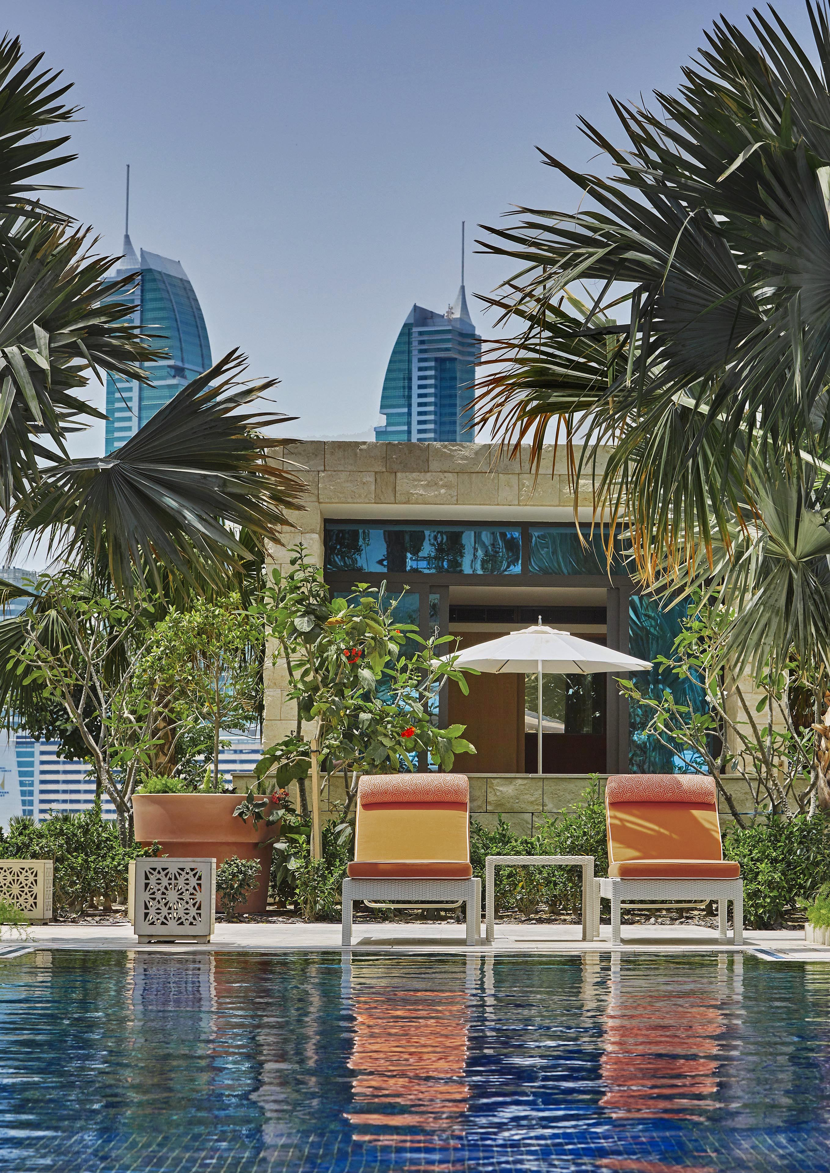 Three luxury hotel lounge chairs by a pool are surrounded by Four Seasons Bahrain palm trees and plants, with a stone building and two modern skyscrapers visible in the background under a clear sky.