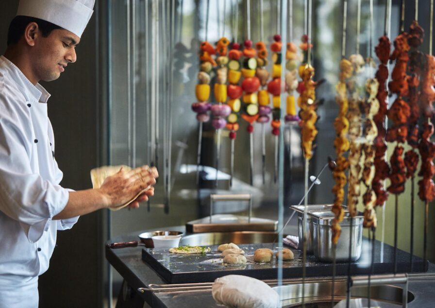A Four Seasons Bahrain lead chef in a white uniform prepares dough at a counter, with skewers of vegetables and meat hanging in the background.