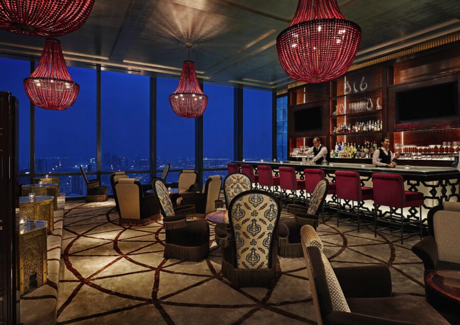 Luxurious Four Seasons Bahrain hotel bar at night with red chandeliers, patterned chairs, a long bar with red stools, two bar service hospitality staff, and large windows overlooking a cityscape.