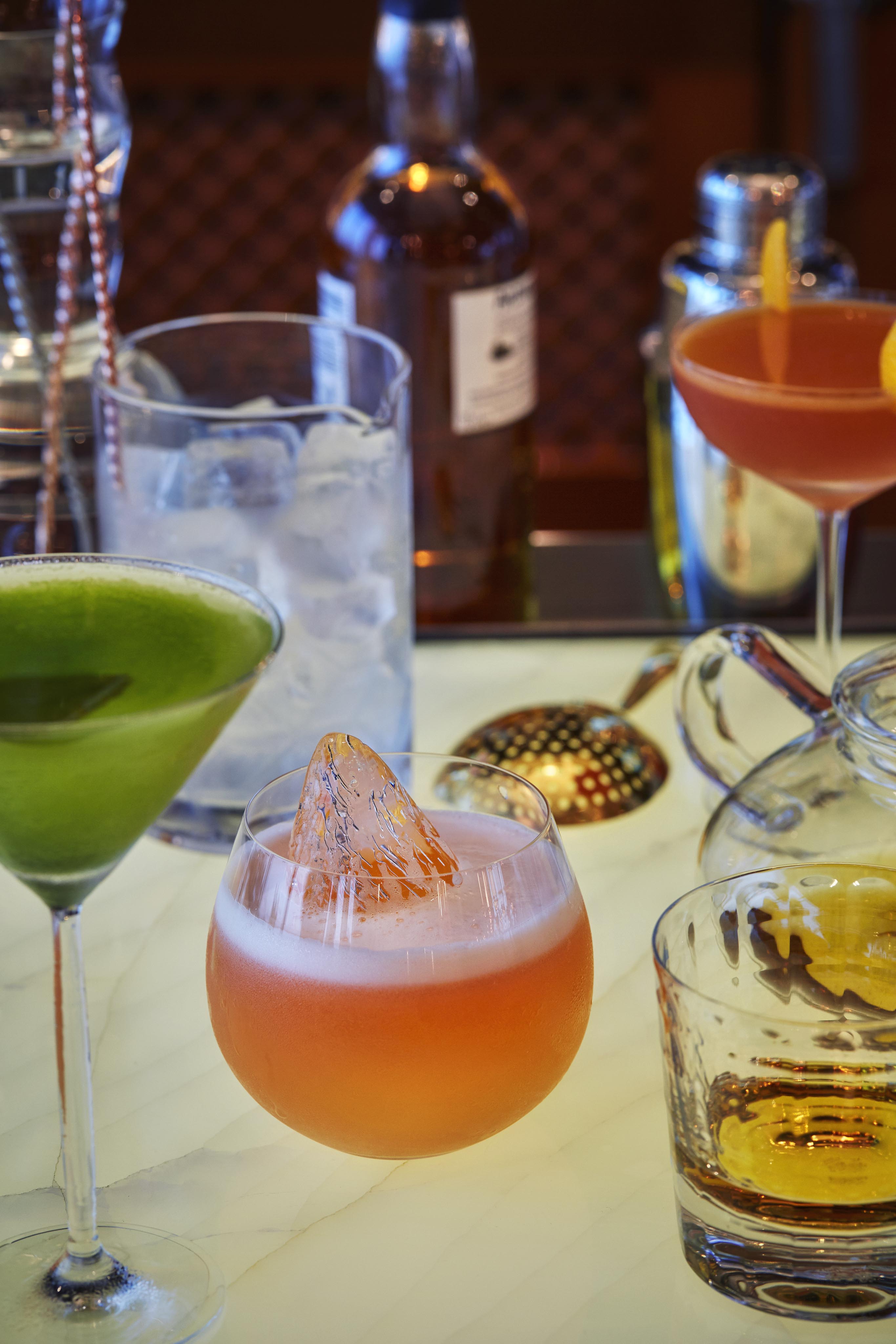 A variety of colorful elevated cocktails in different glasses are displayed on a table, with ice, a bottle, and bar tools in the background—perfectly capturing the vibrant ambiance of luxury hotel and resort photography in the Middle East.