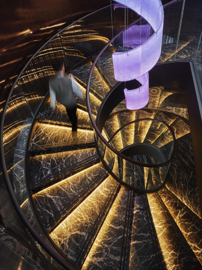Person ascending a spiral staircase of the Four Seasons Bahrain adorned with glowing lights and a purple decorative structure above, set against a dark marble-patterned background.