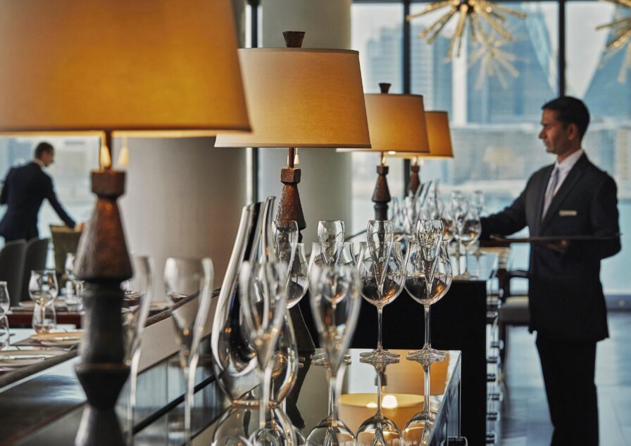 A Four Seasons Bahrain hospitality staff in formal attire arranges glasses on a restaurant table, with lamps, glassware, and city buildings visible in the background.