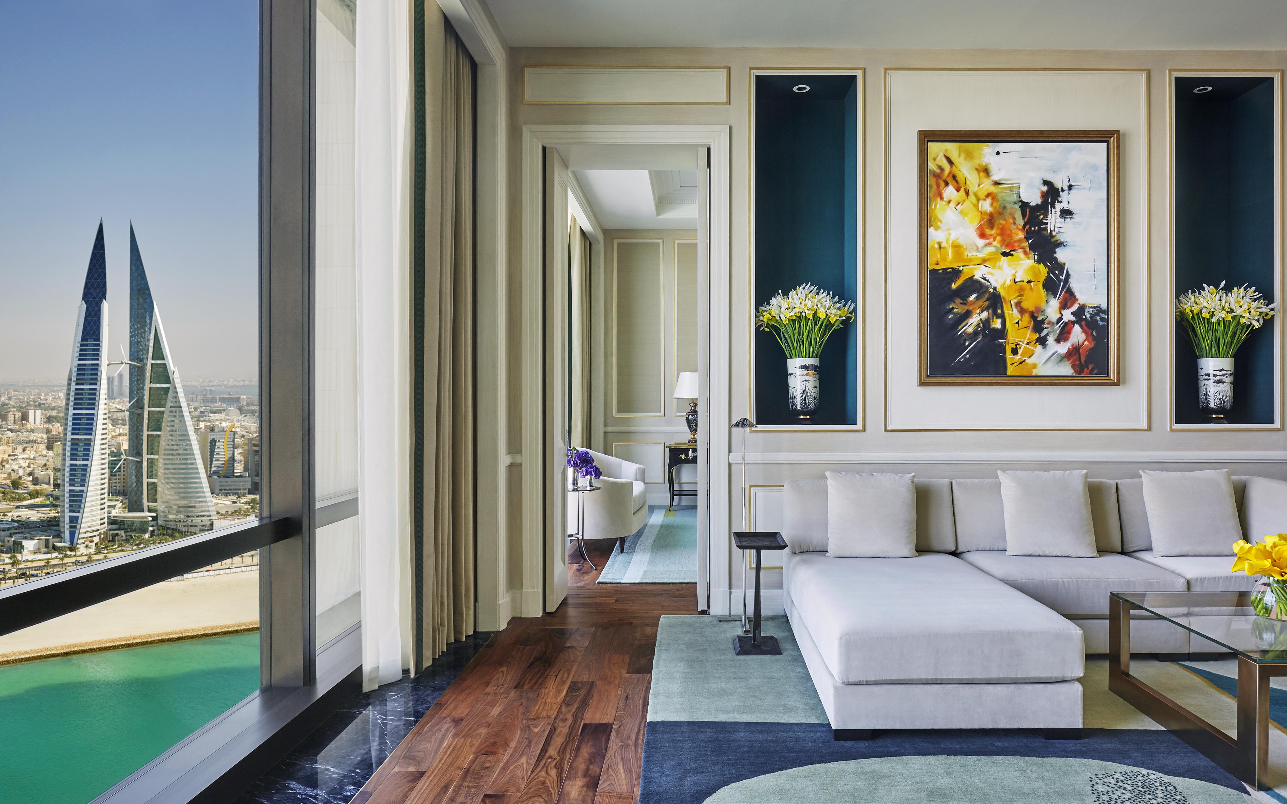Modern luxury Four Seasons Bahrain Bay interior suite with a large white sectional sofa, abstract painting, and floor-to-ceiling window showing a cityscape and iconic twin towers. Vases with flowers adorn the walls.