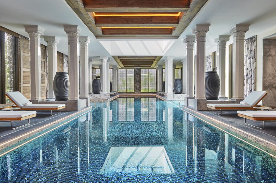 Luxurious Four Seasons Bahrain Bay interior indoor pool with mosaic tiles, surrounded by elegant columns and pristine wooden loungers, under a skylight that floods the space with natural light.