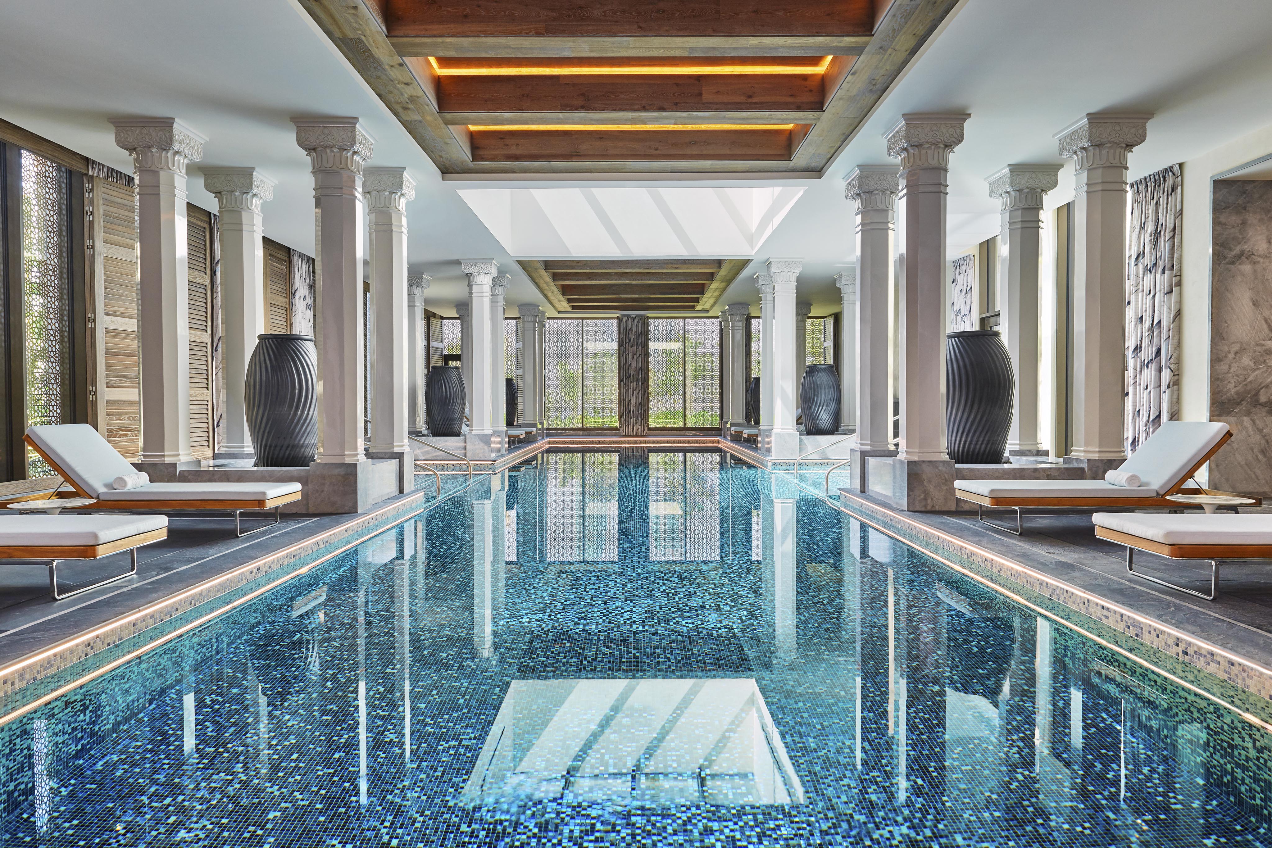 Luxurious Four Seasons Bahrain Bay interior indoor pool with mosaic tiles, surrounded by elegant columns and pristine wooden loungers, under a skylight that floods the space with natural light.