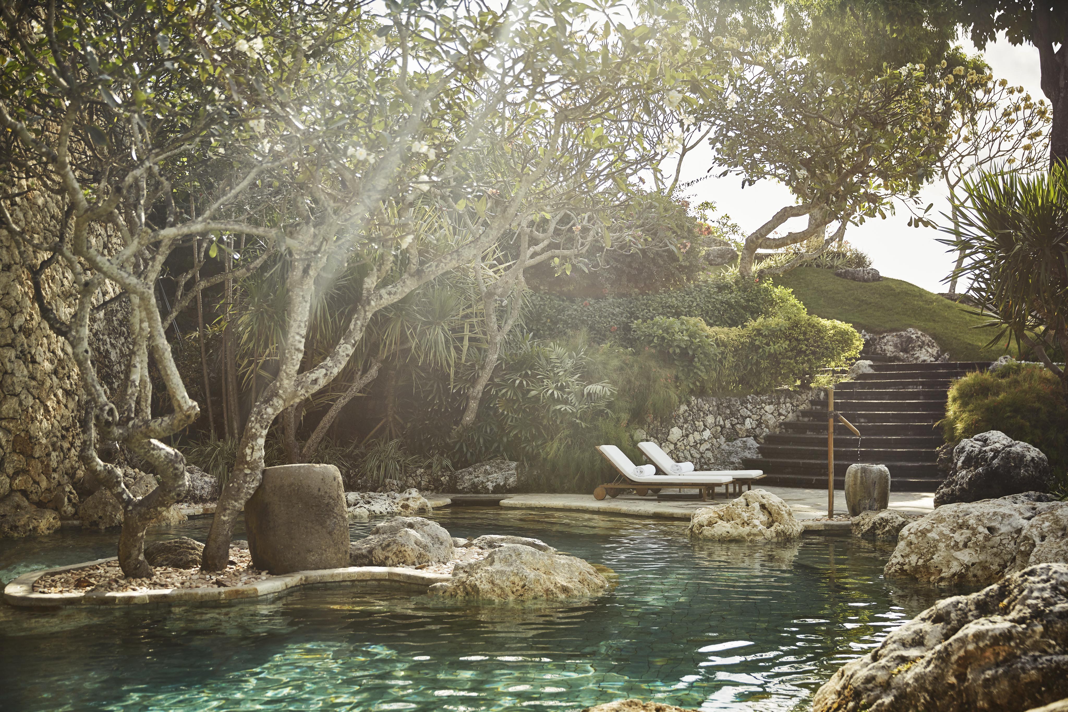 A serene Four Seasons Bali luxury pool surrounded by rocks and lush greenery, with two lounge chairs on a deck in the background. Steps lead up to a higher garden area.