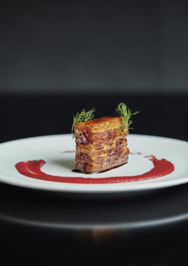 A square piece of seared meat sits on a white plate, garnished with herbs. A swirl of red sauce, inspired by the vibrant flavors of Central America, encircles the meat.