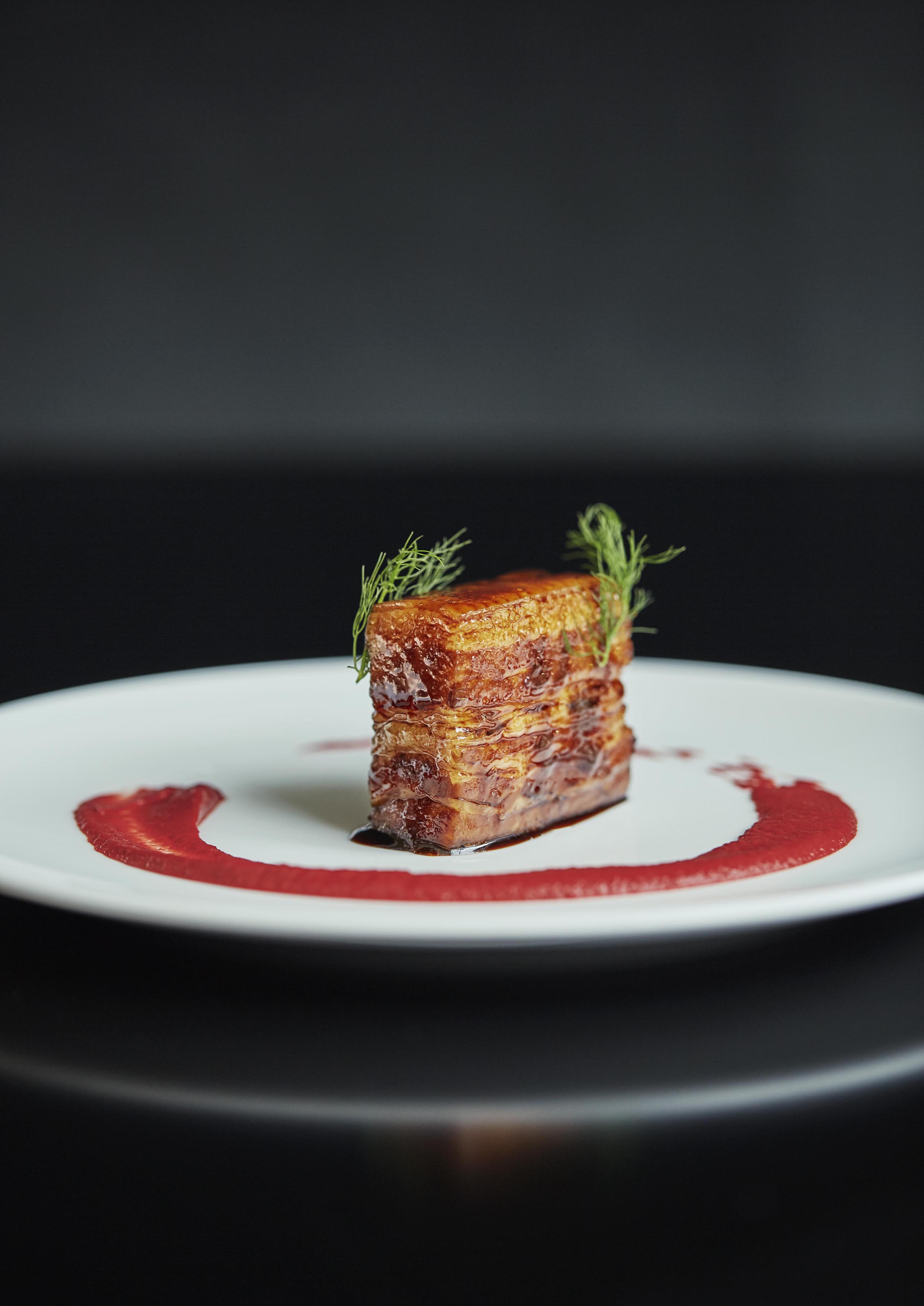 A square piece of seared meat sits on a white plate, garnished with herbs. A swirl of red sauce, inspired by the vibrant flavors of Central America, encircles the meat.