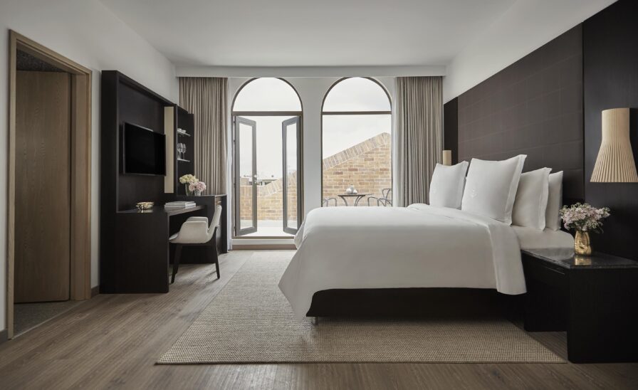Modern hotel room featuring a large bed with a dark headboard, wall-mounted TV, and desk. An open door invites you to a balcony with a stunning city view, offering a touch of South American charm.
