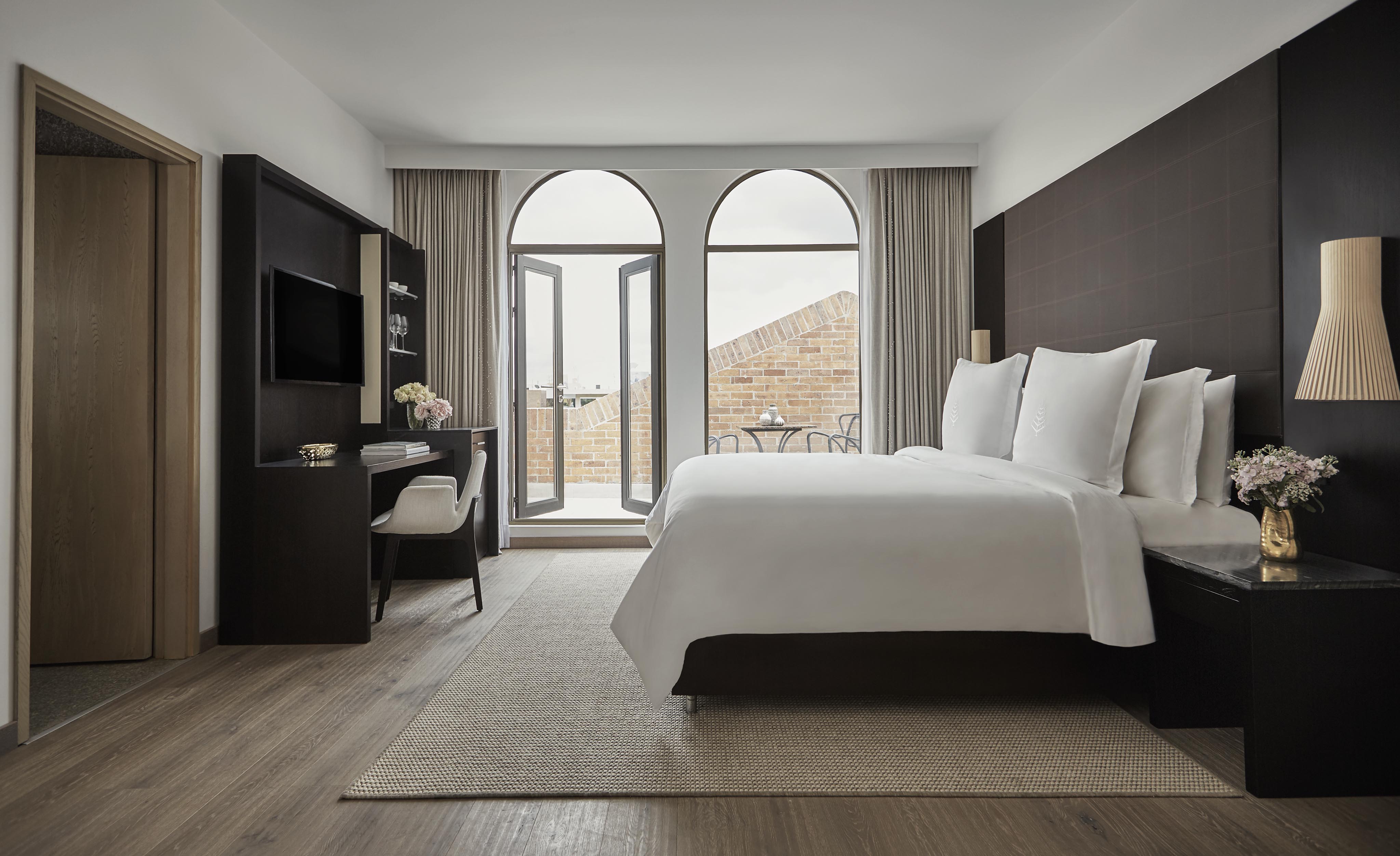Modern hotel room featuring a large bed with a dark headboard, wall-mounted TV, and desk. An open door invites you to a balcony with a stunning city view, offering a touch of South American charm.