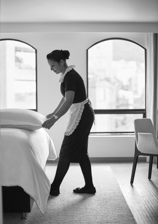 A hotel room attendant in uniform meticulously makes a bed, adjusting the pillow with precision. The room features a chair and large windows offering an urban view reminiscent of vibrant South America. Black and white image.