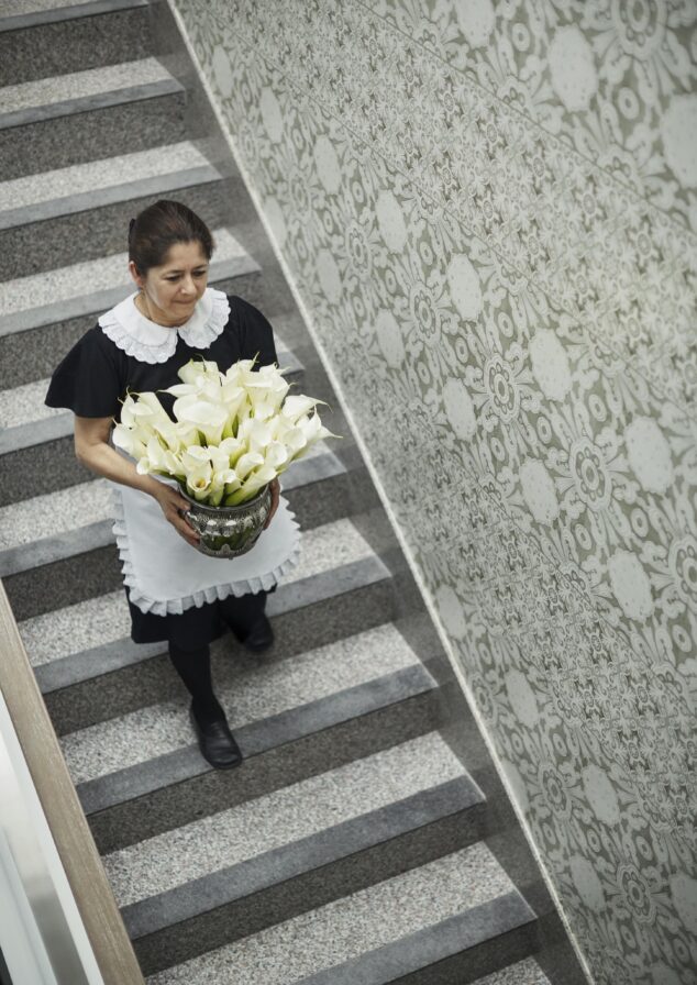 A woman in a uniform gracefully carries a vase of white calla lilies down a staircase adorned with vibrant, patterned wallpaper reminiscent of the vibrant art found throughout Central America.