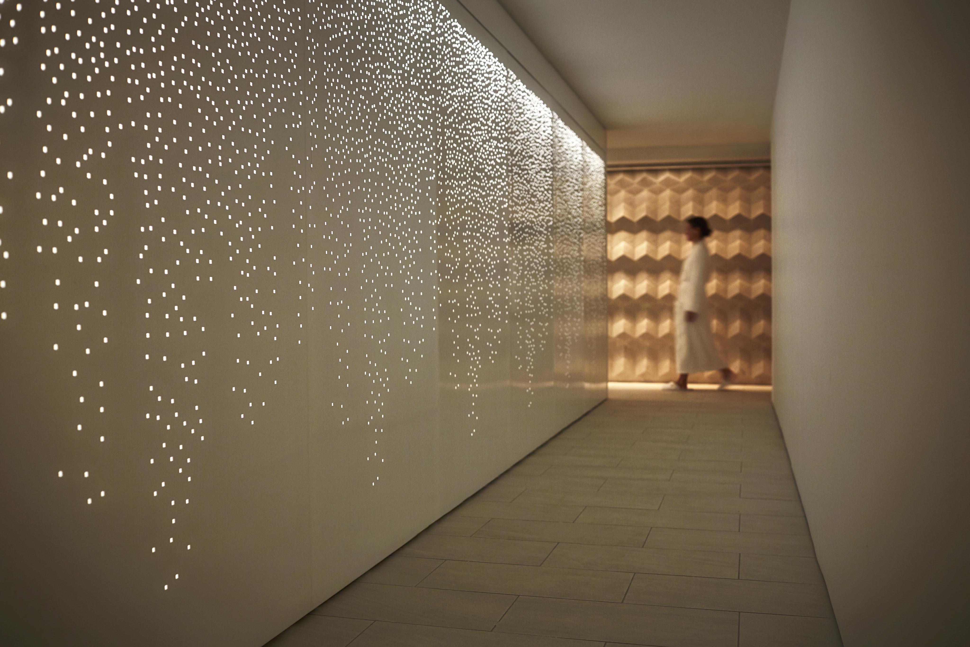 A person in white strolls down a hallway, reminiscent of South America's vibrant architecture, with a wall of small, glowing holes on one side and a beautifully patterned wall at the end.