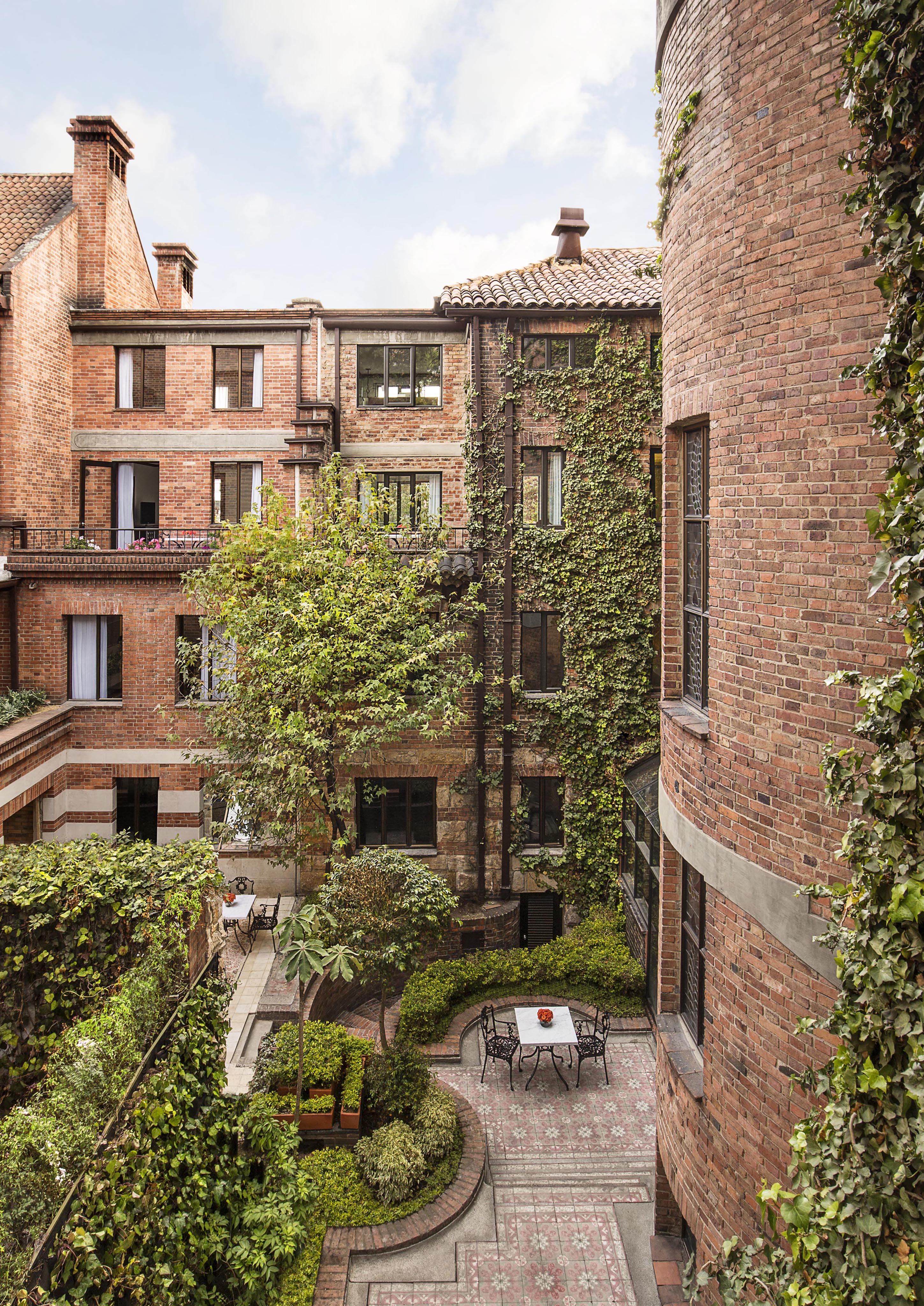 A brick building exterior of the Four Seasons Bogota hotel and resort with ivy-covered walls and multiple windows surrounds a small courtyard reminiscent of Central America. The courtyard features a tree, potted plants, and a table with two chairs on a charming brick patio.
