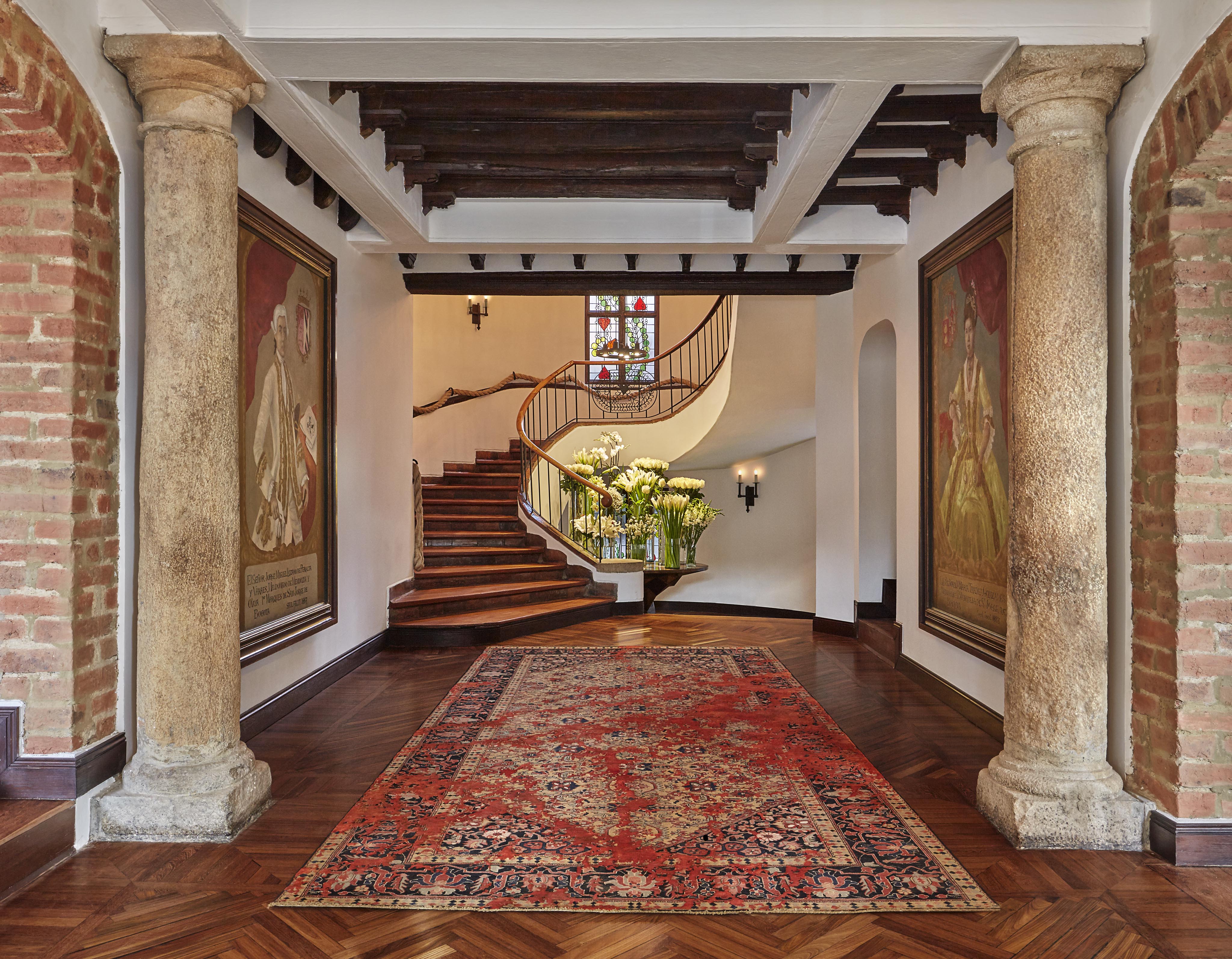 Elegant foyer of Four Seasons Bogota hotel lobby with brick pillars, large tapestry, and red patterned rug evokes a touch of Central American charm. A curved wooden staircase ascends to the upper floor, while a floral arrangement sits gracefully under a stained-glass window.