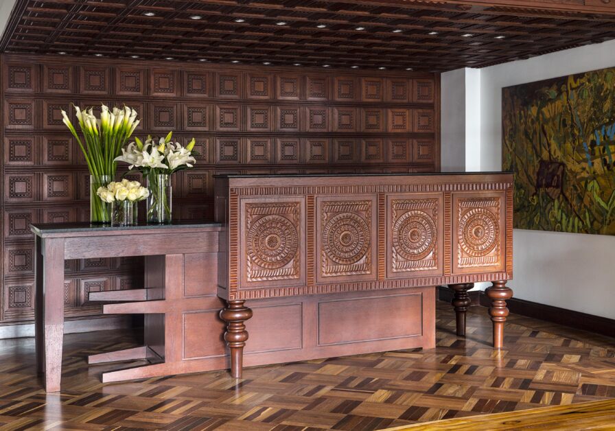 A wooden reception desk of Four Seasons Bogota with ornate carvings, flanked by two vases filled with vibrant flowers, sits against a textured wood wall and parquet floor. On the right, a colorful abstract painting adds flair reminiscent of South American artistry.