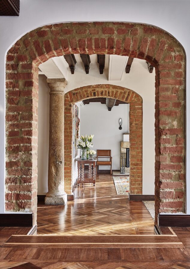 Architectural capture of Four Seasons Bogota luxury hotel lobby with brick archways and a stone pillar exuding a classic charm reminiscent of South America. The wooden flooring leads the eye to a room where a chair, table, and vase of flowers create an inviting nook.