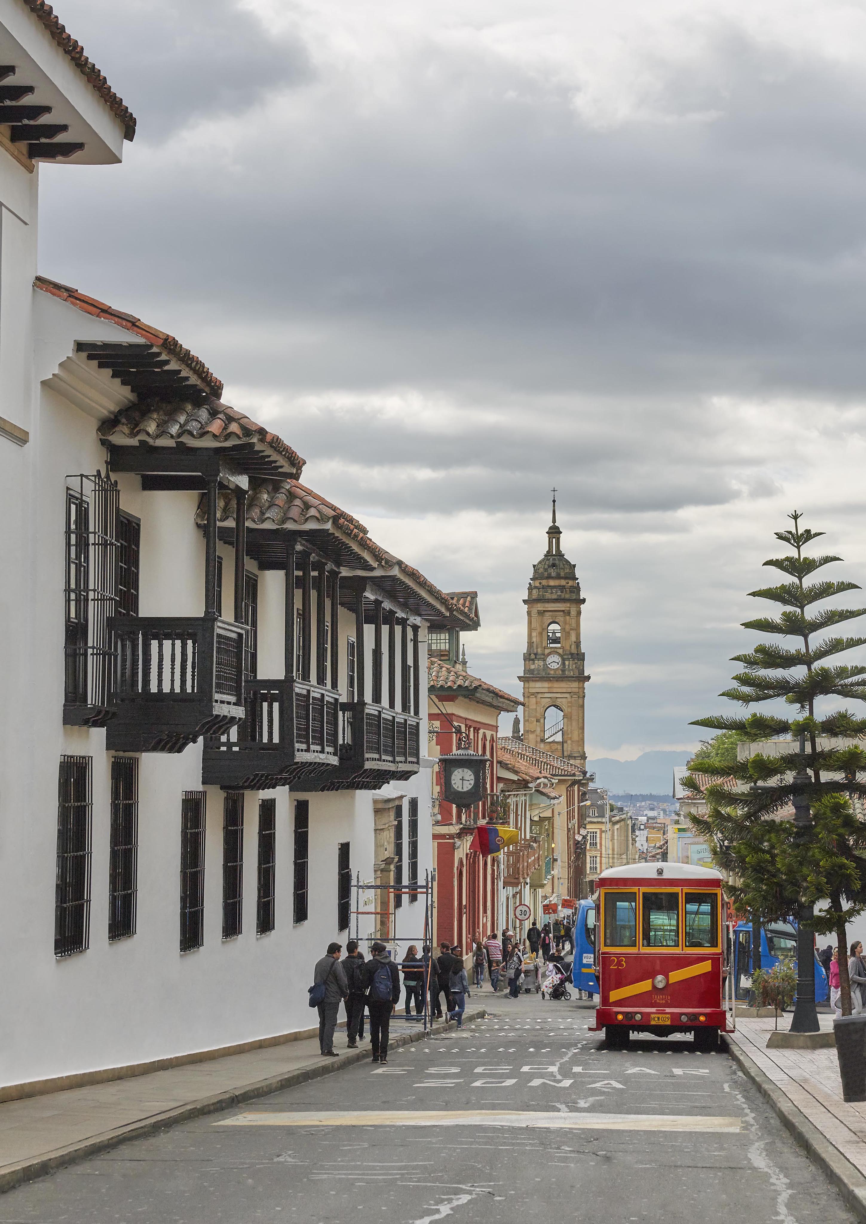 A street scene with people and a red bus in a historic city area of Central America. Colonial-style buildings line the street, and a clock tower is visible under a cloudy sky.