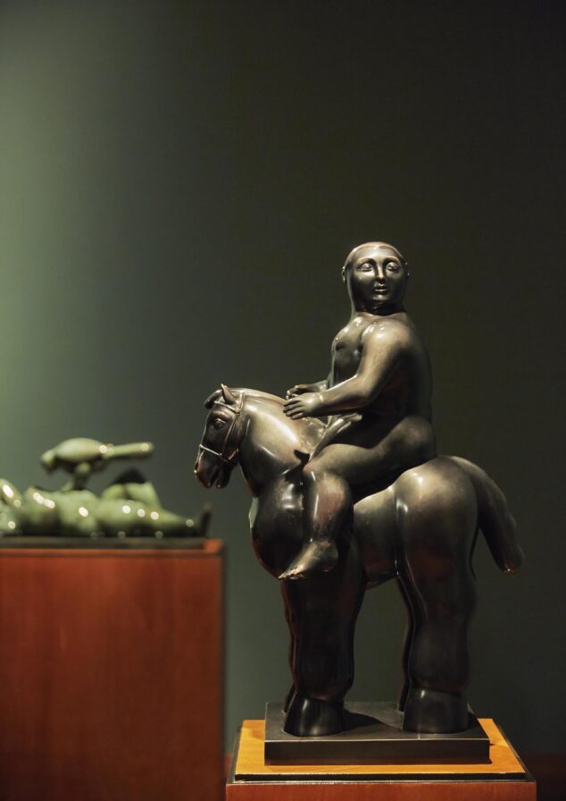 In a dimly lit gallery, a bronze sculpture of a person on a horse commands attention, reminiscent of equestrian traditions in South America. In the background, another majestic horse sculpture complements the scene.