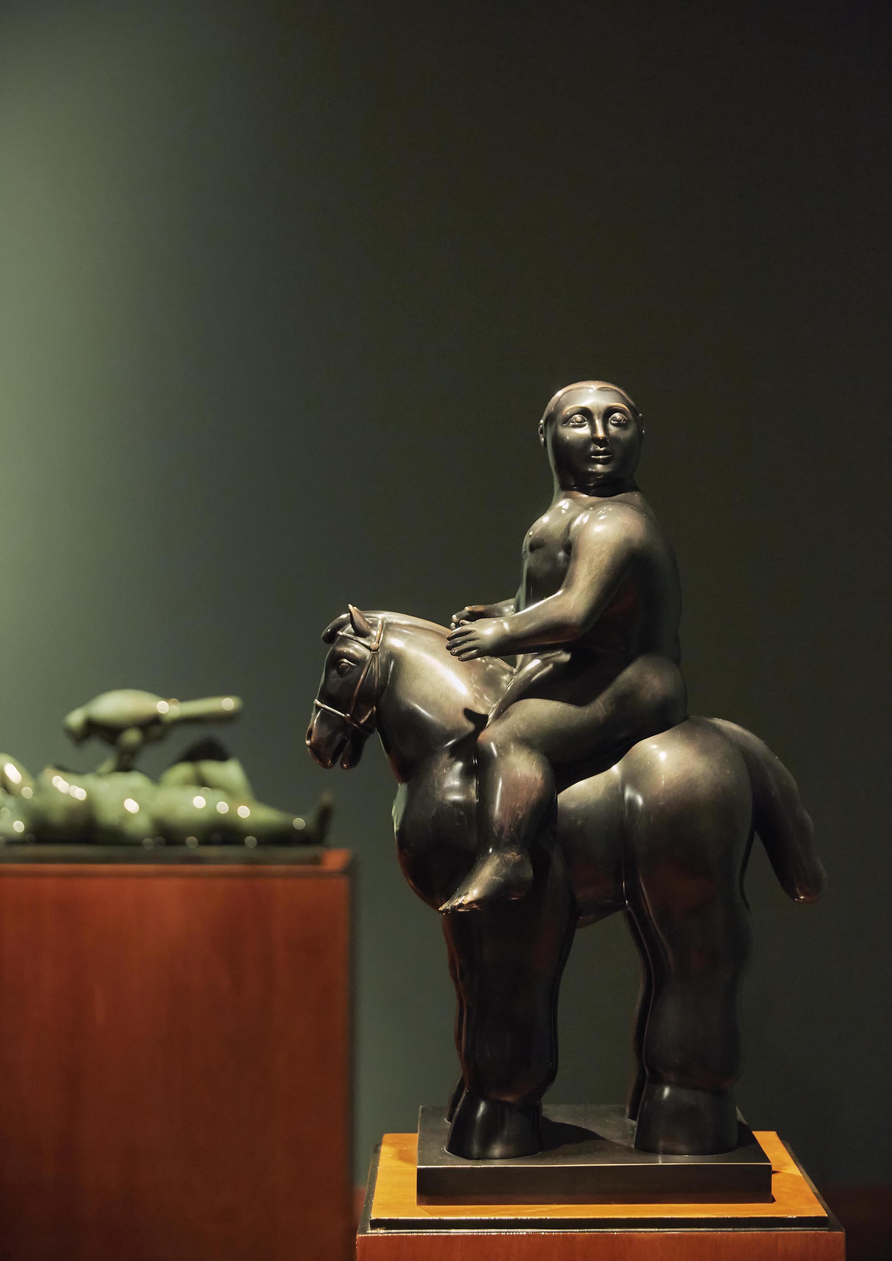In a dimly lit gallery, a bronze sculpture of a person on a horse commands attention, reminiscent of equestrian traditions in South America. In the background, another majestic horse sculpture complements the scene.