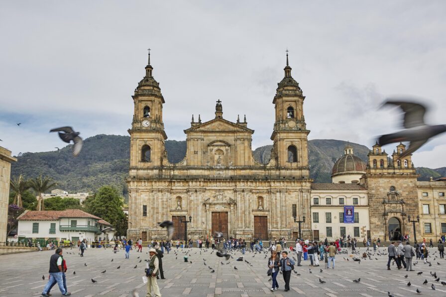 A large historical of Plaza de Bolivar cathedral with two towers stands at a bustling plaza in Central America, where many people and pigeons create lively scenes. Mountains are visible in the background under a cloudy sky.