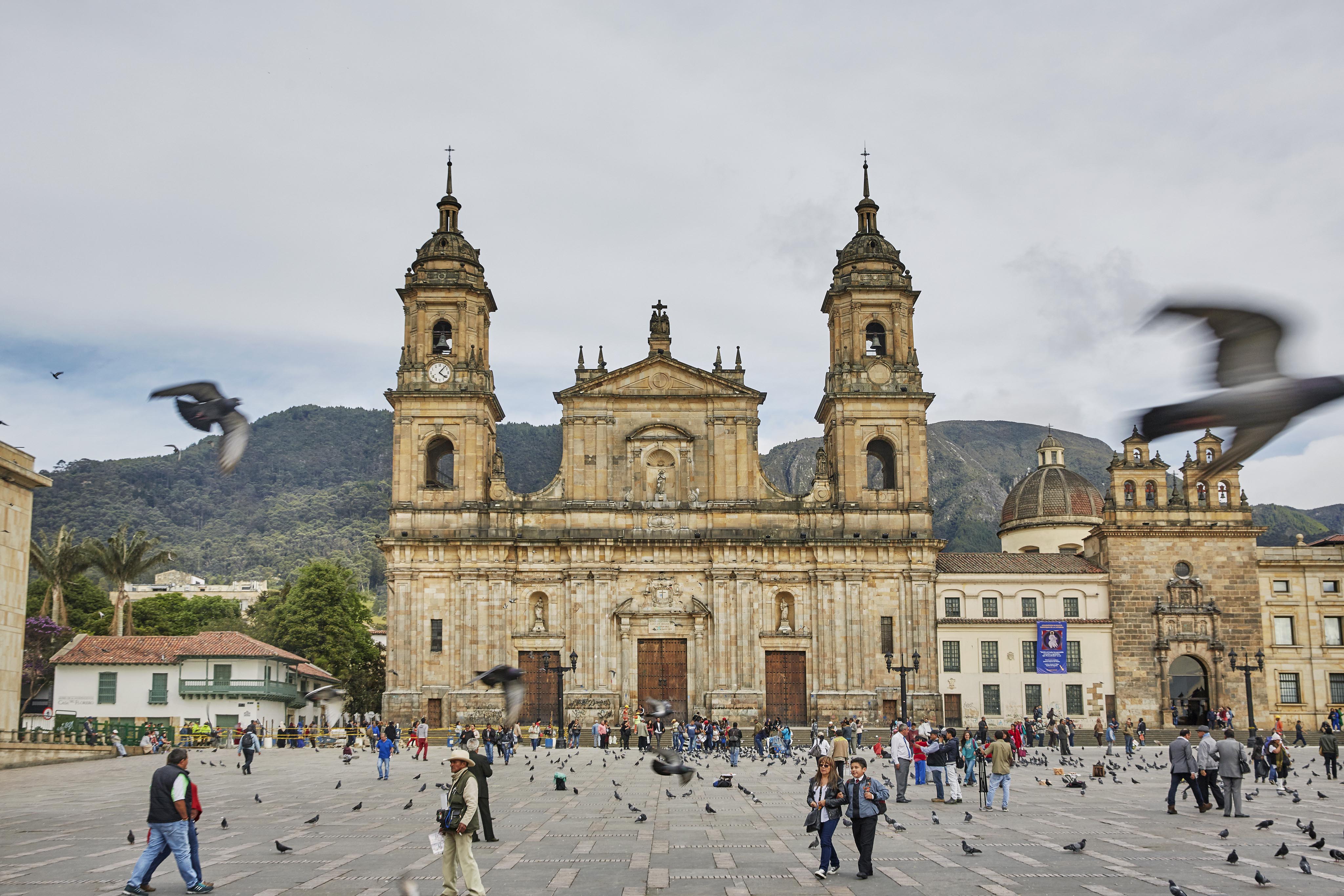 A large historical of Plaza de Bolivar cathedral with two towers stands at a bustling plaza in Central America, where many people and pigeons create lively scenes. Mountains are visible in the background under a cloudy sky.