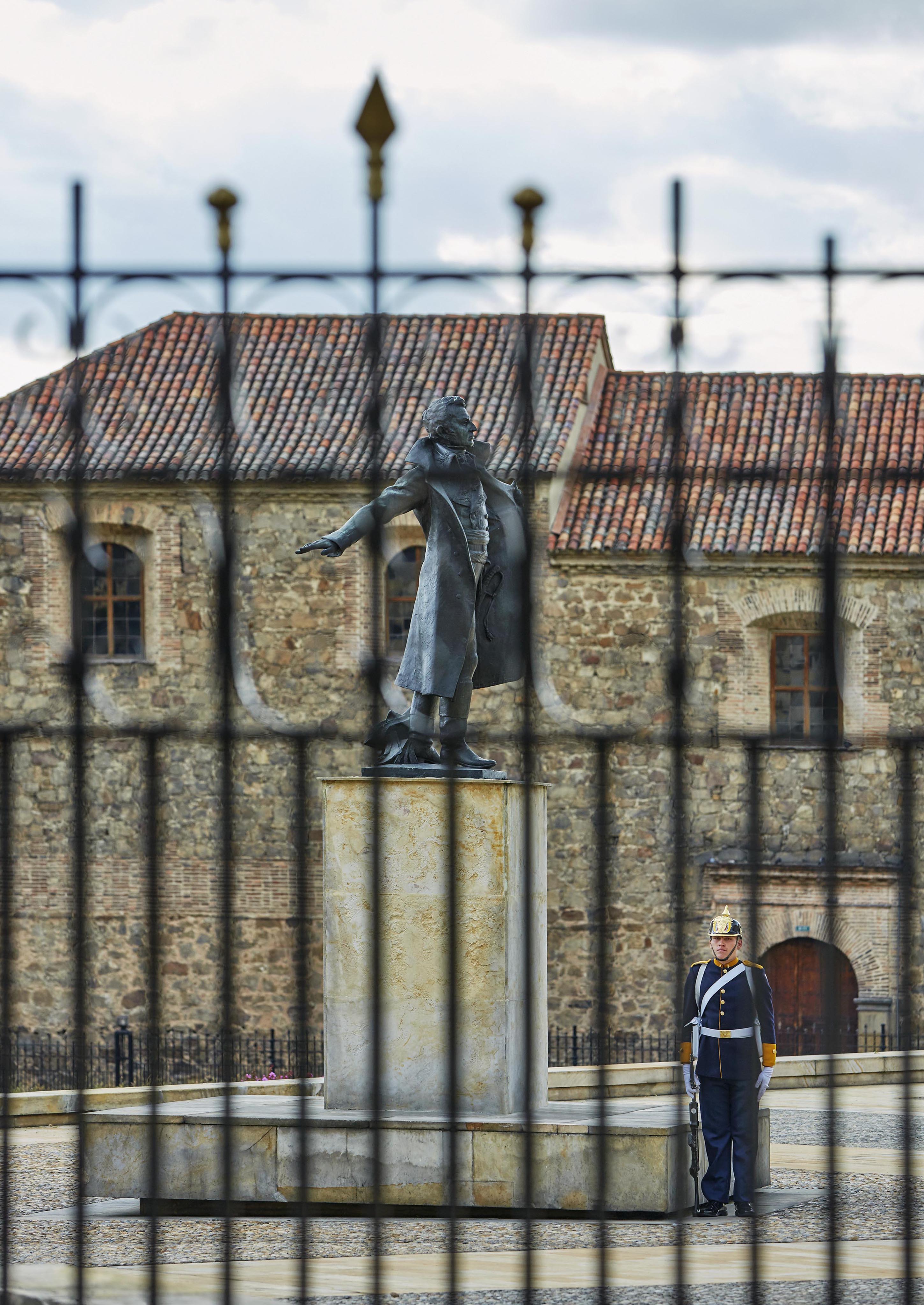A statue on a pedestal stands behind a metal fence, watched over by a uniformed guard to the right. The scene is reminiscent of South America's historic charm, with a stone building featuring a red-tiled roof in the background.