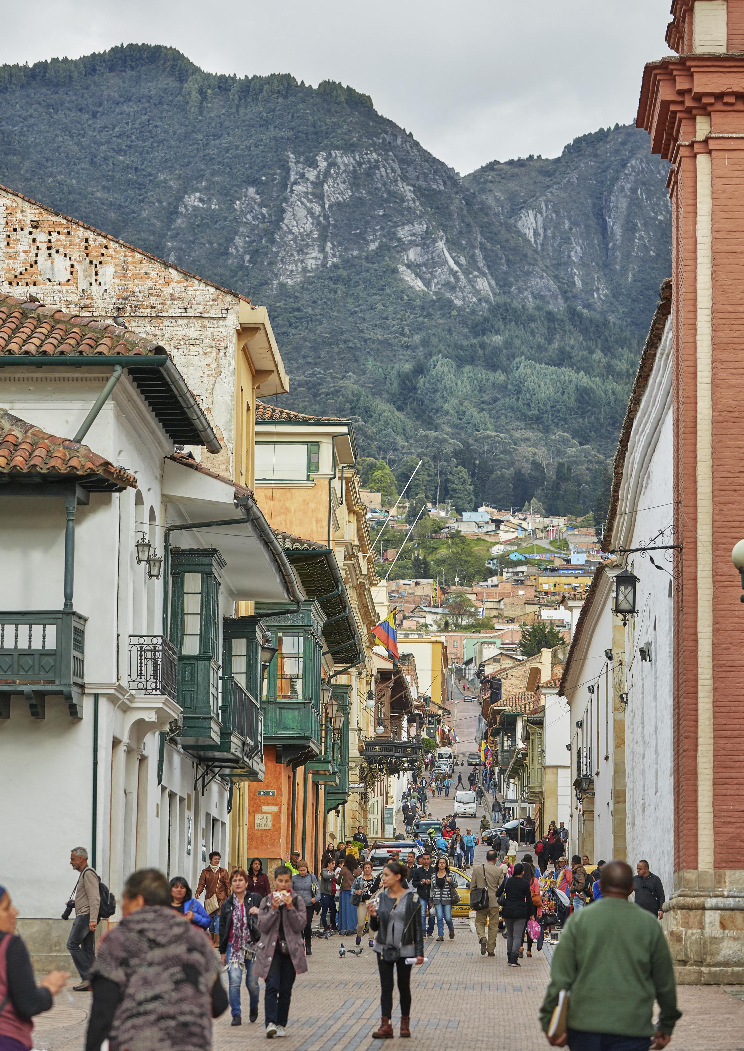 A bustling street in a historic South American town with colorful buildings, numerous pedestrians, and majestic mountains in the background.