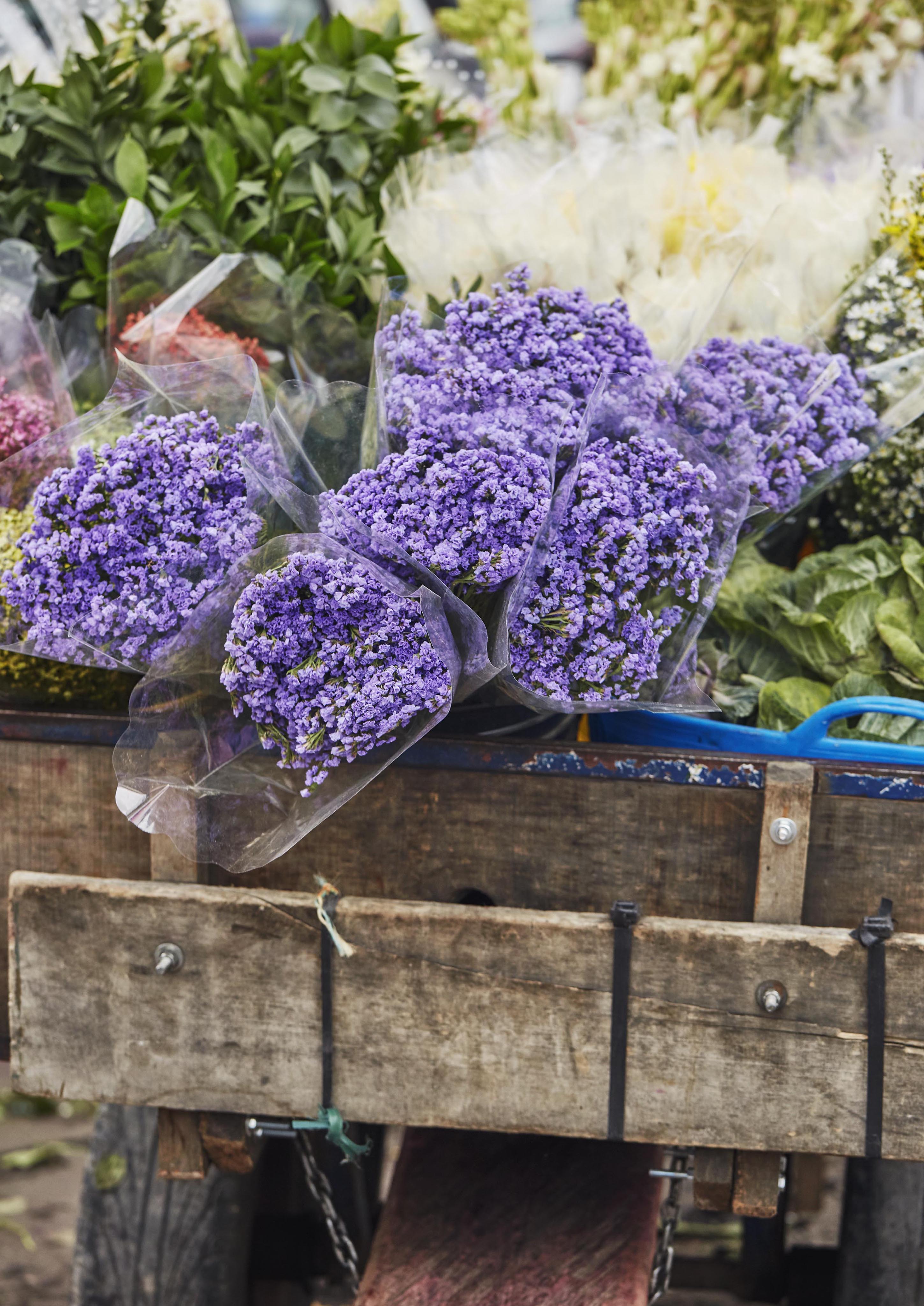 Bouquets of purple flowers wrapped in plastic are displayed on a wooden cart at an outdoor Bogota market, reminiscent of vibrant Latin American markets, with additional greenery and flowers in the background.