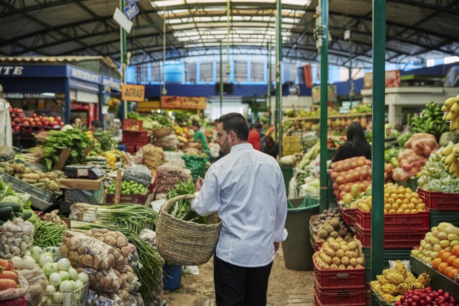 A person in a white shirt carries a basket through a bustling South American market, with vibrant fruits and vegetables displayed in colorful piles under a covered roof.