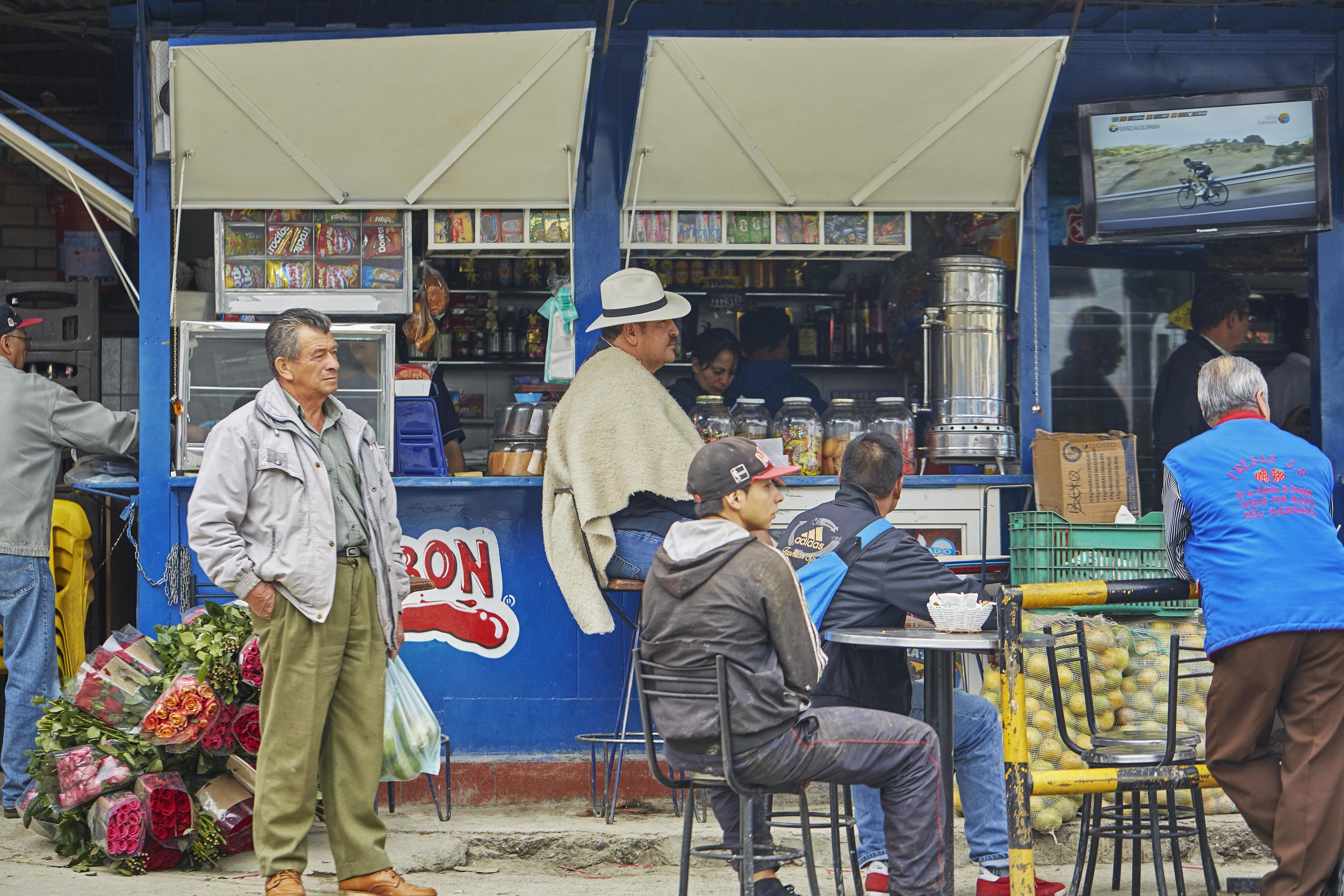 People gather outside a small blue market stall reminiscent of a vibrant South American scene. A man sells food and drinks, with tables of potatoes and flowers nearby. A wall-mounted TV shows a cycling race, capturing the lively atmosphere.