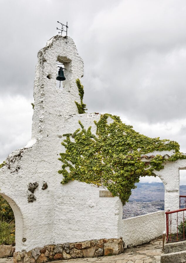 A white stone structure within destination town of Monserrate with a bell, partially covered in green ivy, stands under a cloudy sky. Through its archway, one can glimpse the distant allure of Central America's lush landscapes or perhaps a South American city's vibrant silhouette.
