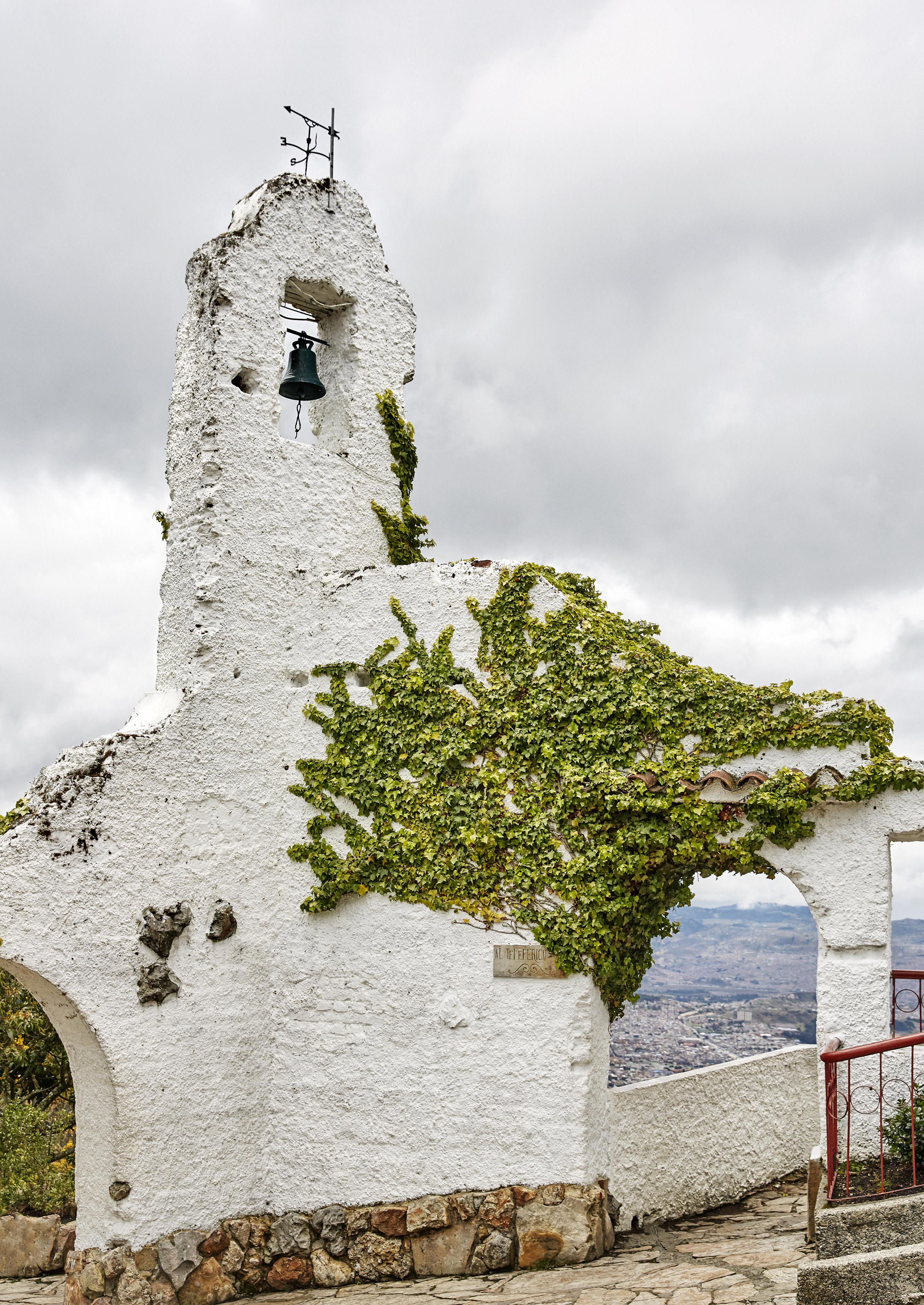 A white stone structure within destination town of Monserrate with a bell, partially covered in green ivy, stands under a cloudy sky. Through its archway, one can glimpse the distant allure of Central America's lush landscapes or perhaps a South American city's vibrant silhouette.