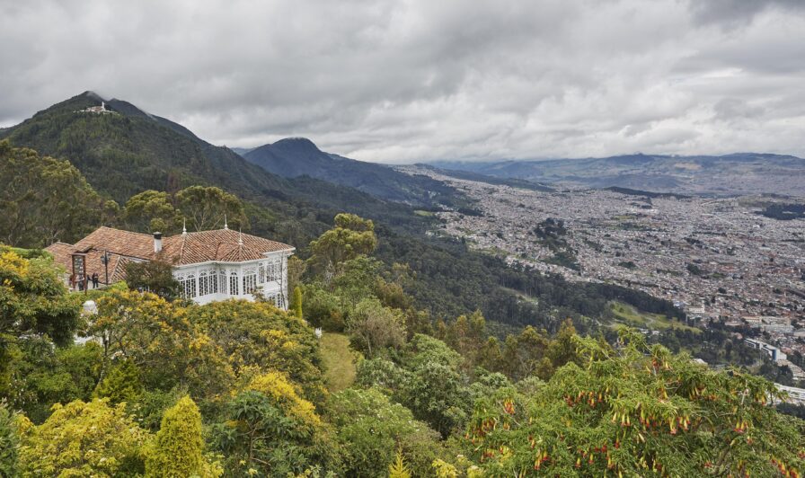Hillside view of a destination city Monserrate, with a white house nestled among lush greenery and mountains under a cloudy sky.