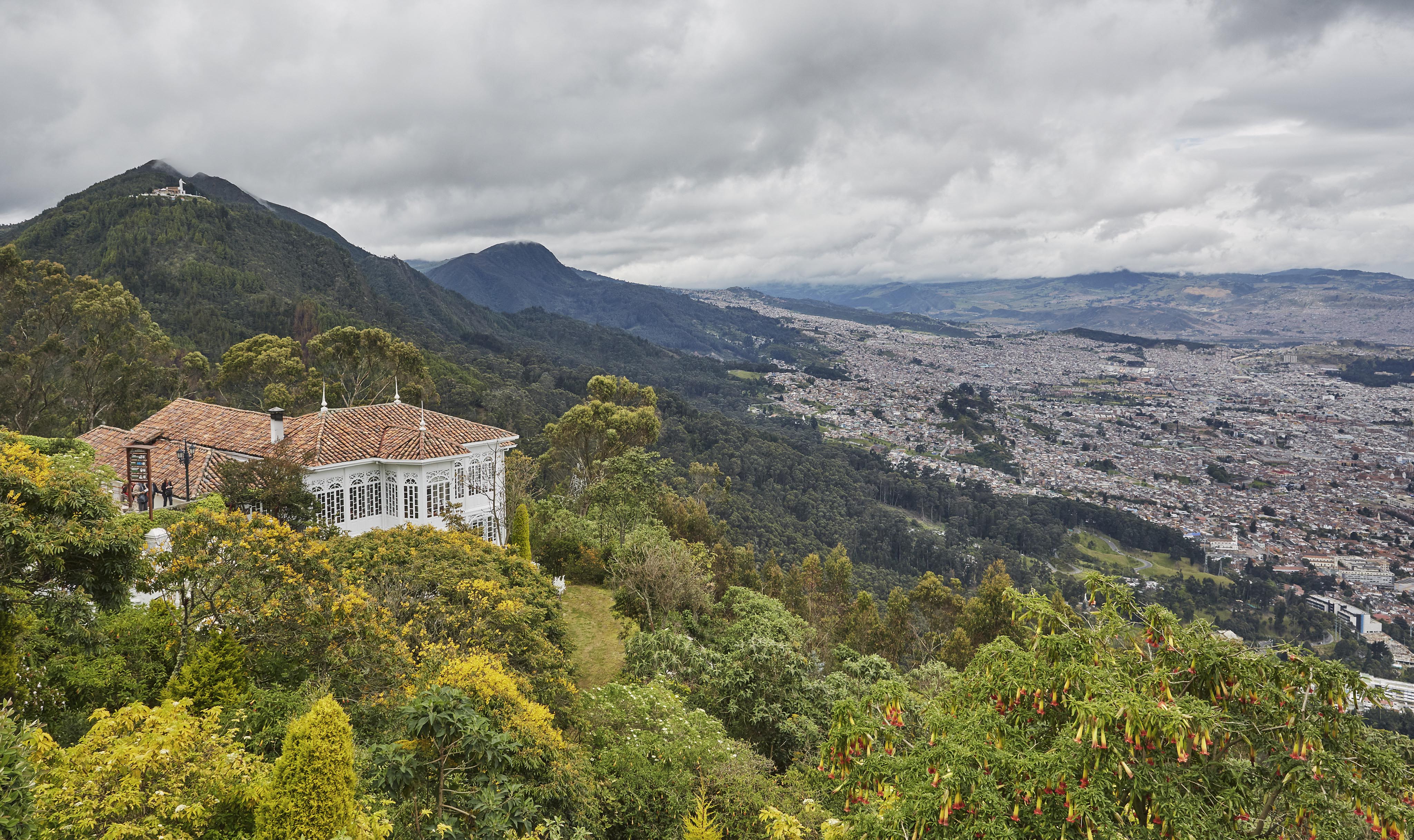Hillside view of a destination city Monserrate, with a white house nestled among lush greenery and mountains under a cloudy sky.