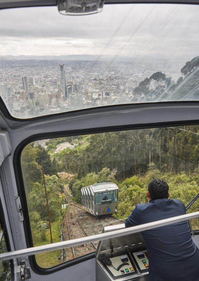 A person in a cable car overlooks a vibrant destination cuty of Monserrate, typical of Central America, with another car approaching on a track, surrounded by lush greenery.